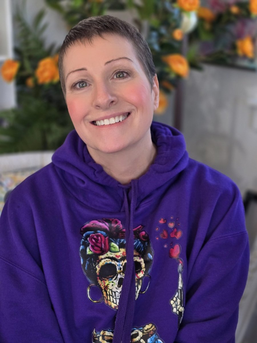 Person with short hair smiling, wearing a purple hoodie with a colorful skull design, in a setting with flowers in the background.