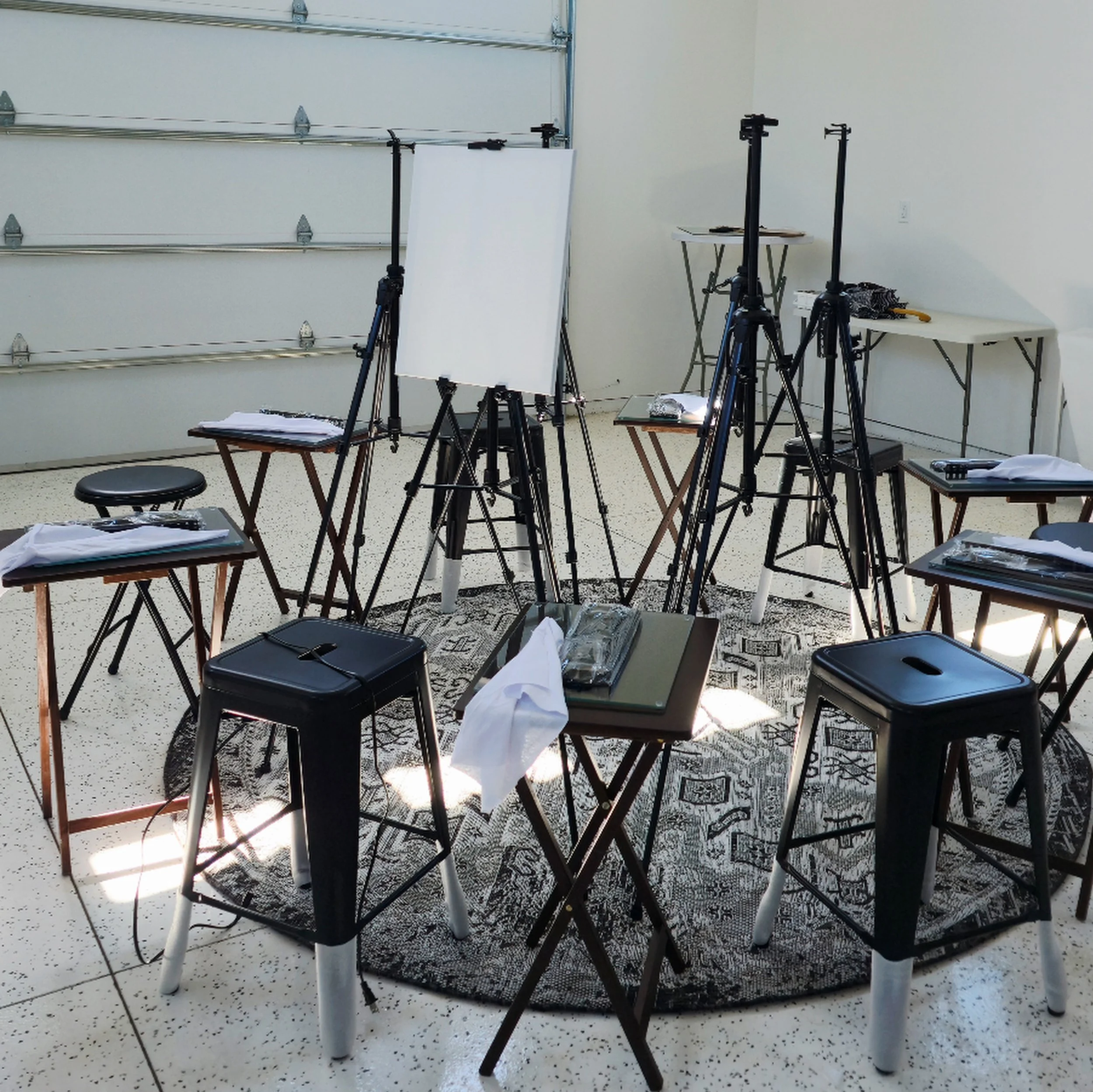 A photography studio setup with tripods, a whiteboard, and tables with papers and equipment in a room with white walls and speckled floor.