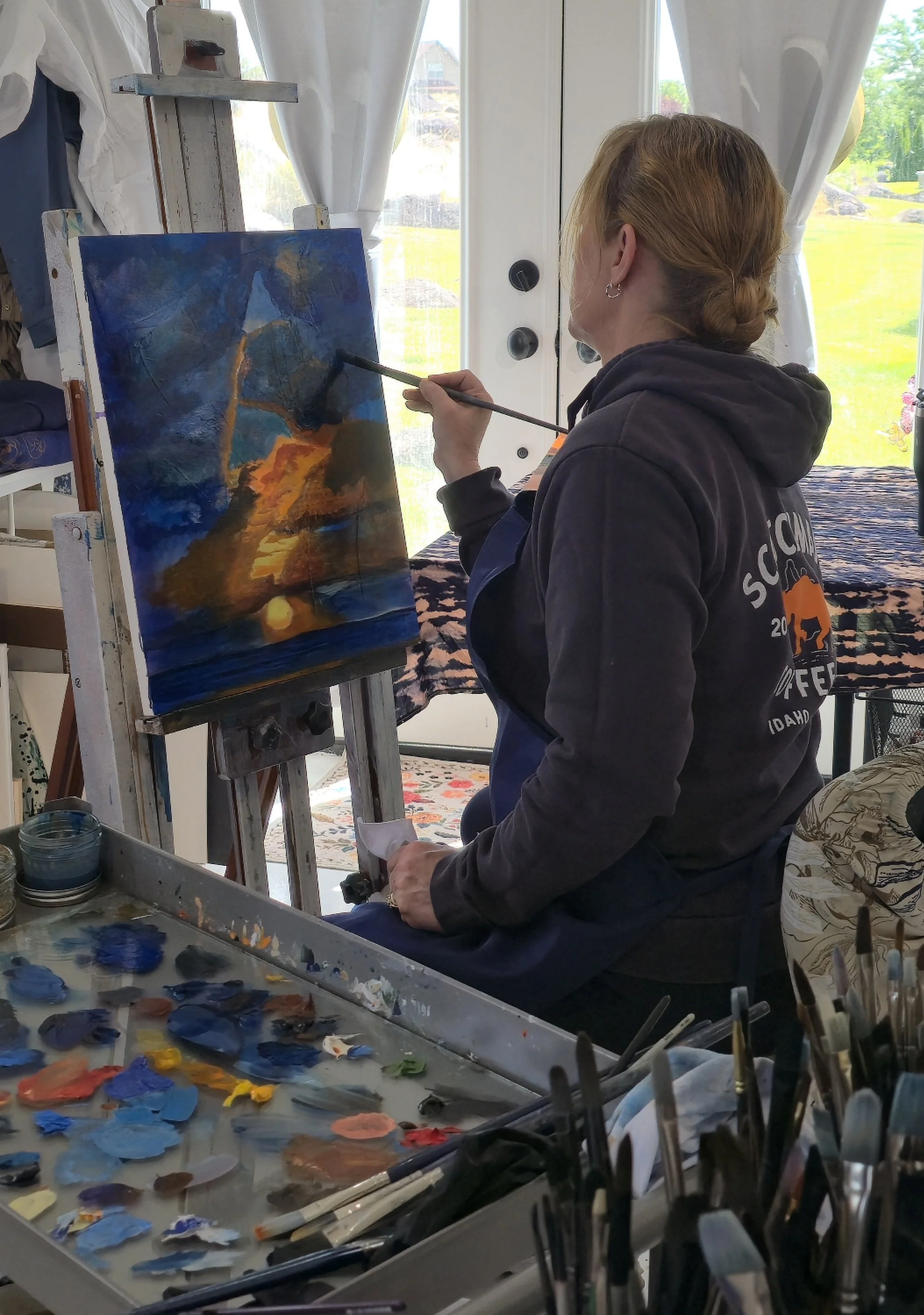 Person with red hair painting an abstract landscape with blue, orange, and yellow colors on a canvas, surrounded by painting supplies in a bright room with large windows showing a green outdoor scene.