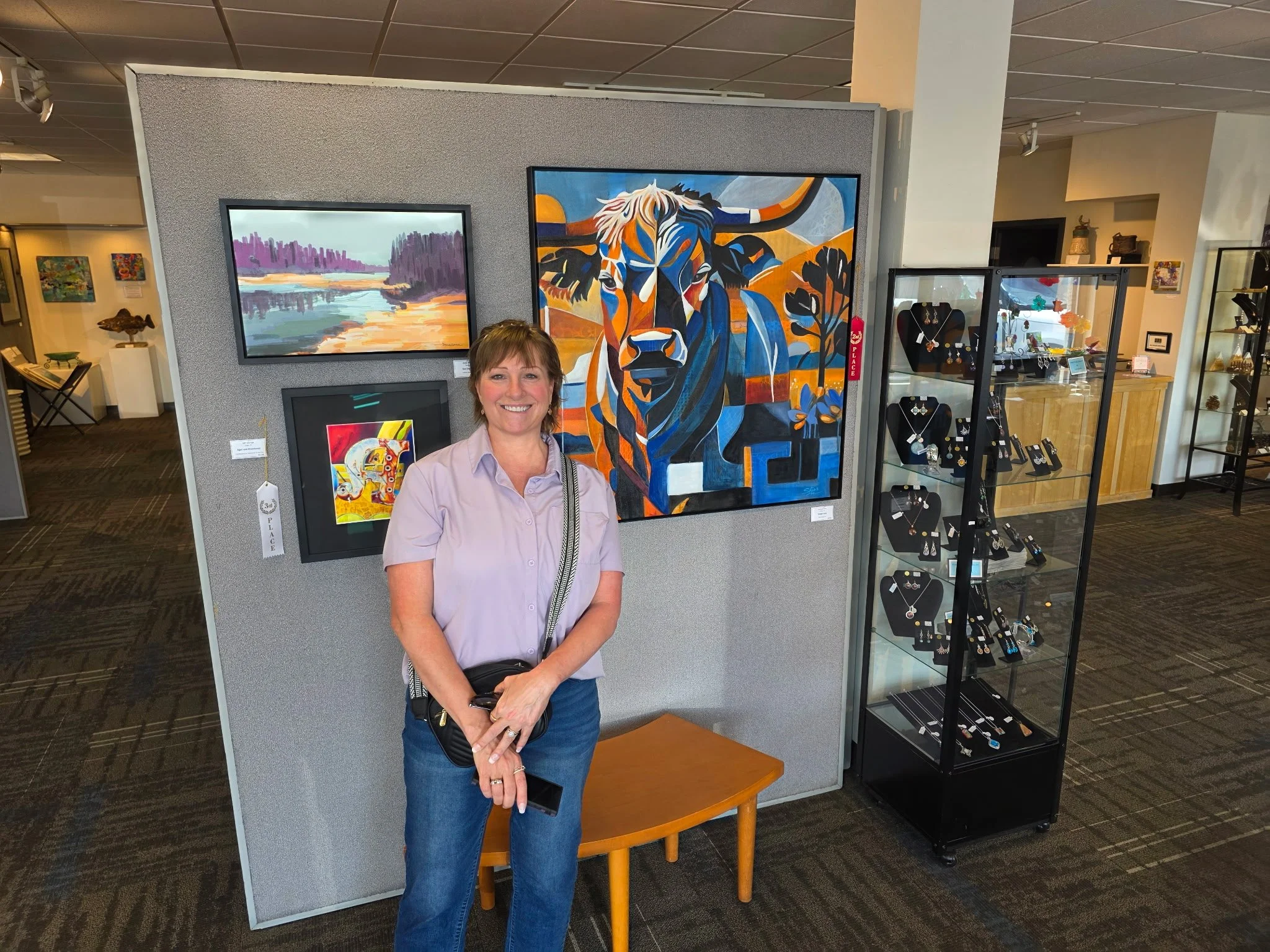 A woman with short hair, wearing a light purple shirt and jeans, smiling while standing in an art gallery. Behind her are colorful paintings, one of a river scene and another of a stylized cow. To her right are jewelry displays in a glass case.