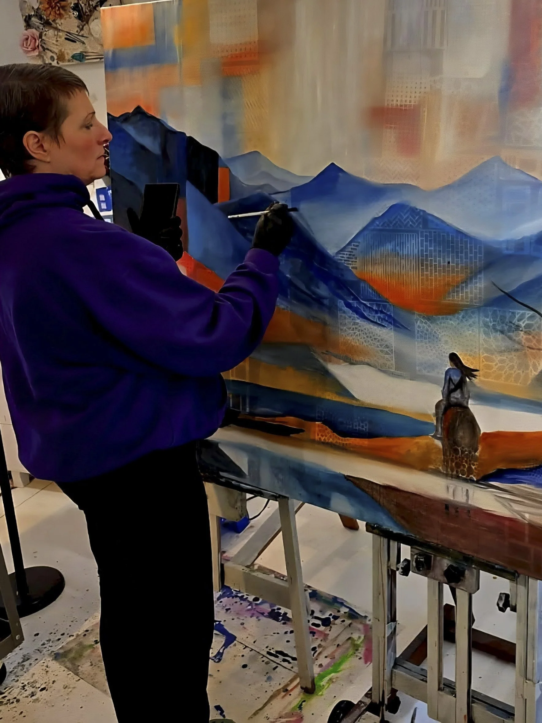 An artist painting a mural of blue mountains and a giraffe on a large canvas in an art studio. The artist, wearing a purple hoodie and black gloves, is standing and working with a paintbrush.