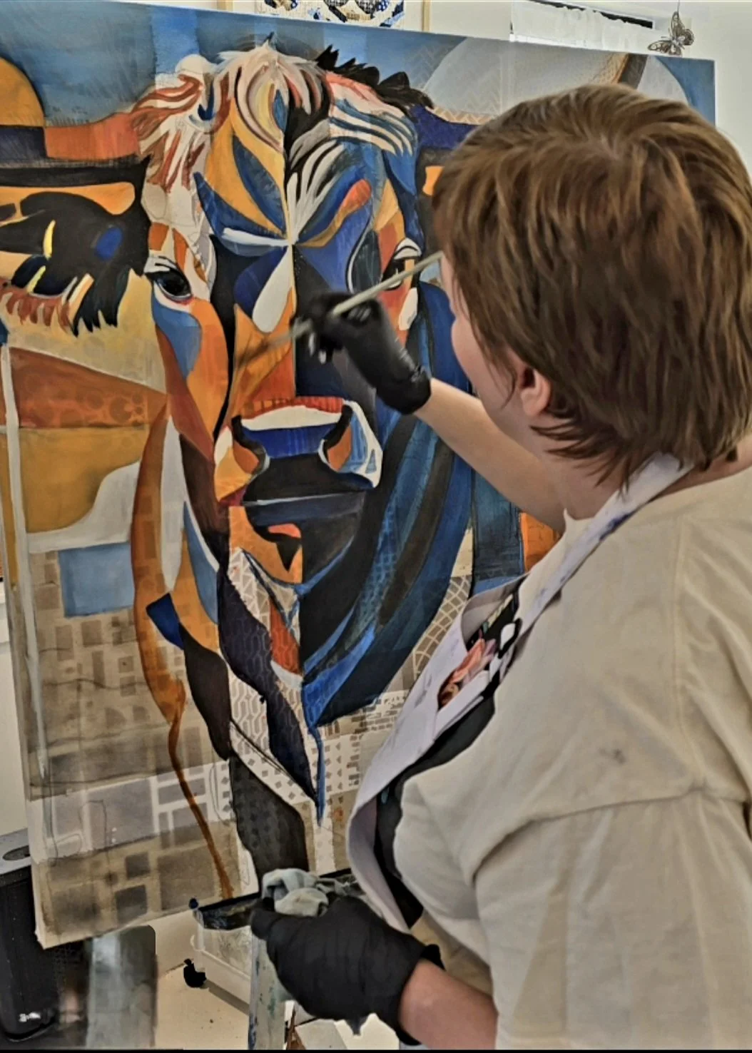 An artist is painting a colorful, abstract portrait of a wolf's face on a large canvas. The artist is wearing a beige apron and black gloves.