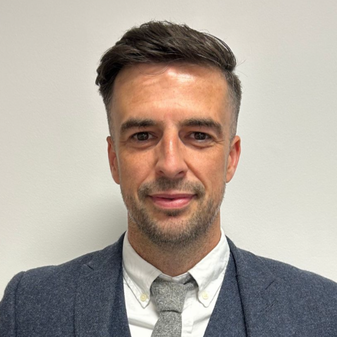 A man with brown hair and a beard wearing a dark gray blazer, white shirt, and gray tie, standing against a plain white wall.