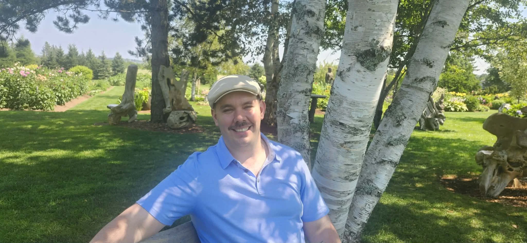 A smiling man in a light blue shirt and beige cap sitting outdoors next to a white birch tree, with green grass, trees, and colorful flowers in the background.