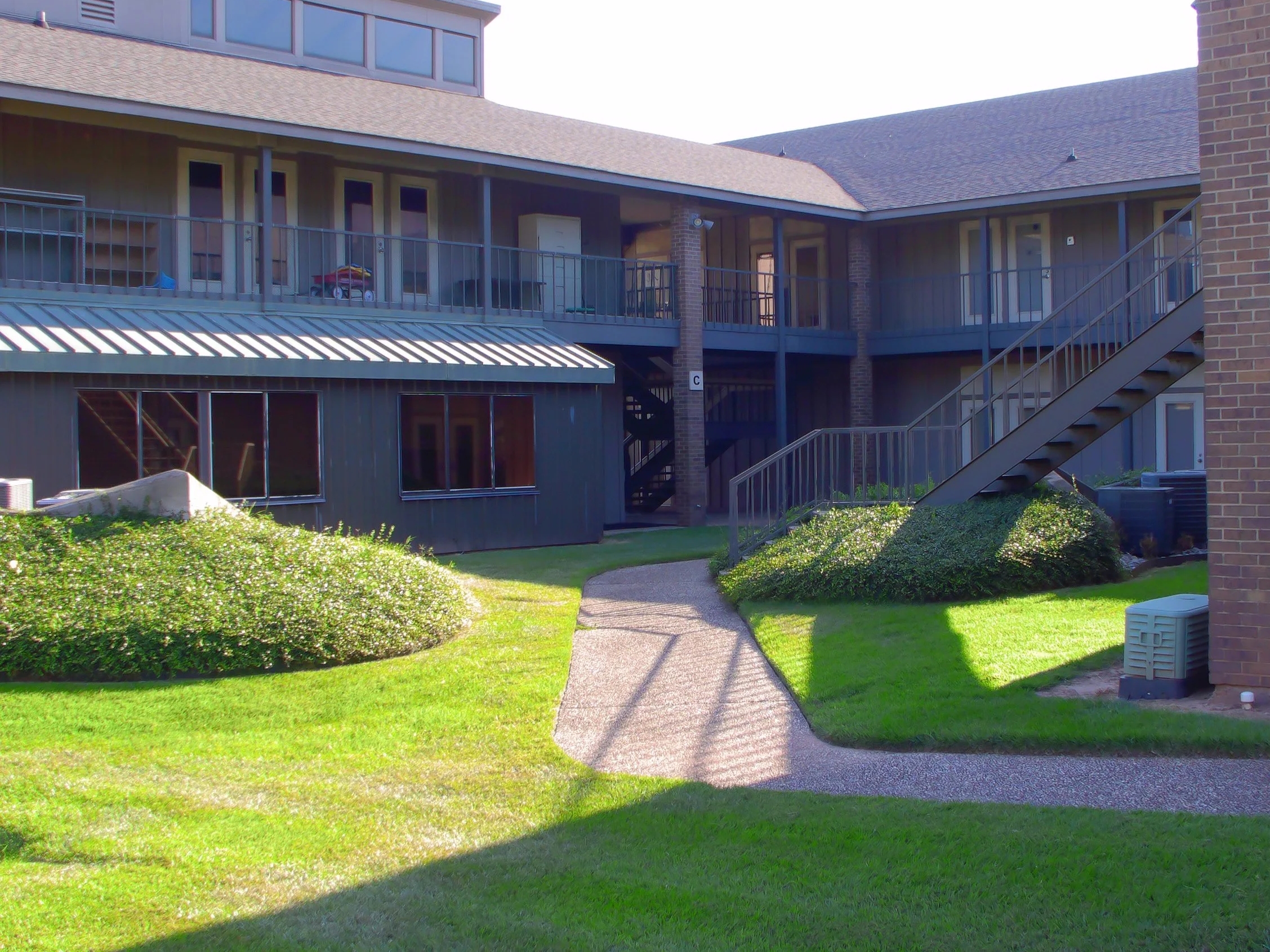 Exterior view of an apartment building with a second-floor balcony, stairs, and landscaped lawn with bushes and a pathway.
