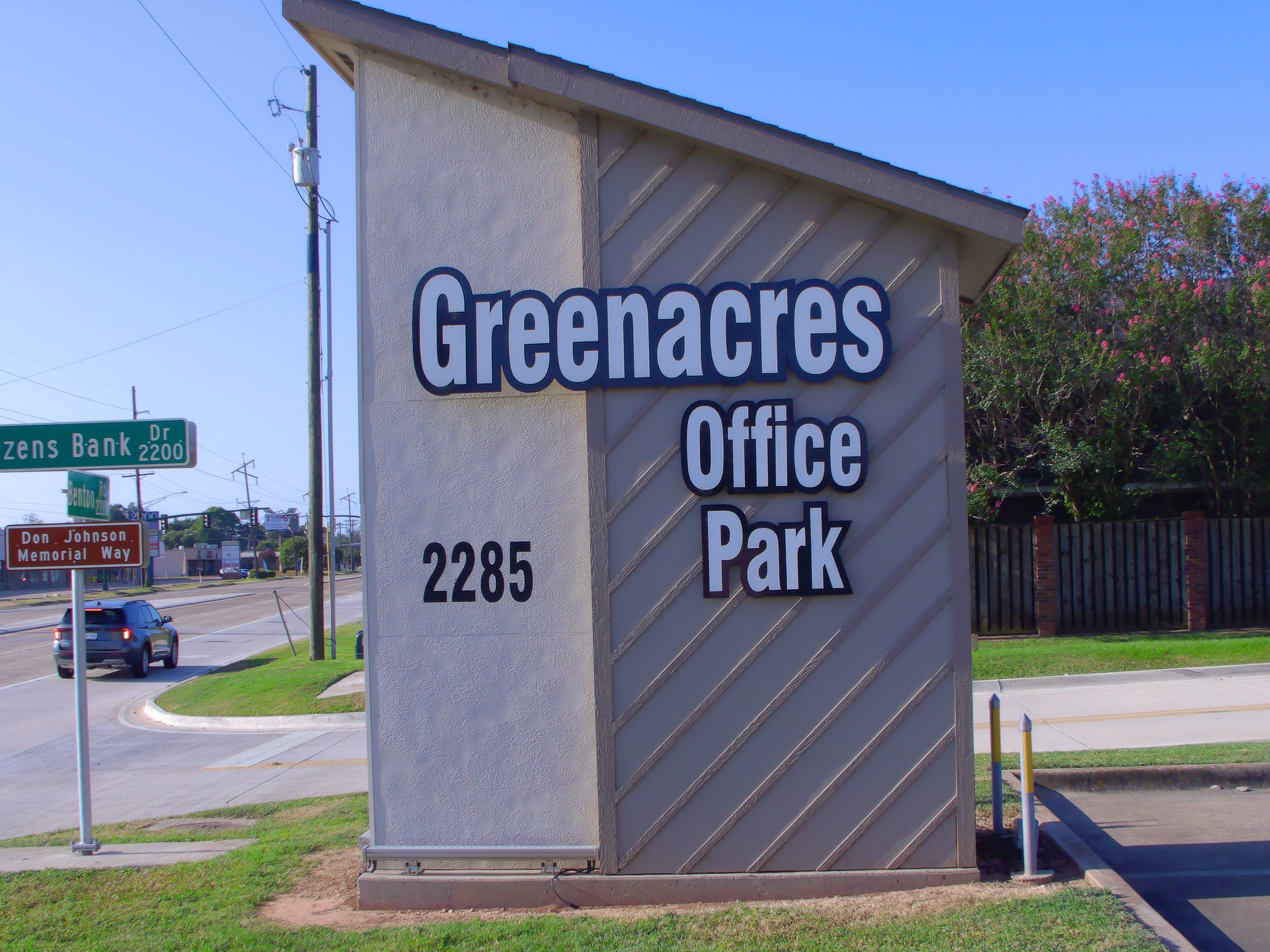 Street corner with street signs, sidewalk, and a large sign for Greenacres office and park. Cars are on the road, with trees and power lines in the background.
