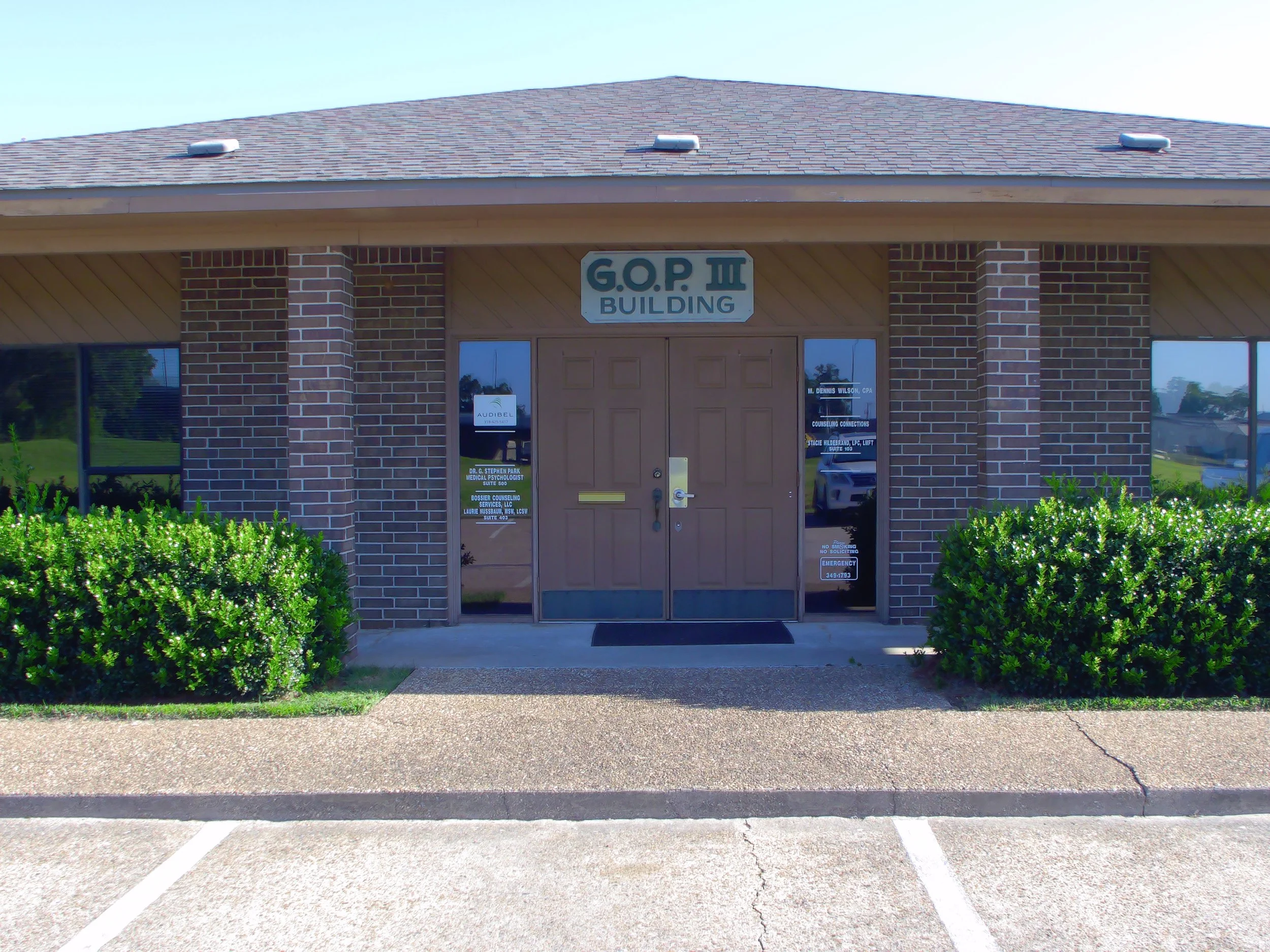 Front view of a brick office building with a sign reading 'G.O.P III Building' above the entrance. Green bushes line the sidewalk in front, with parking spaces empty in the foreground.