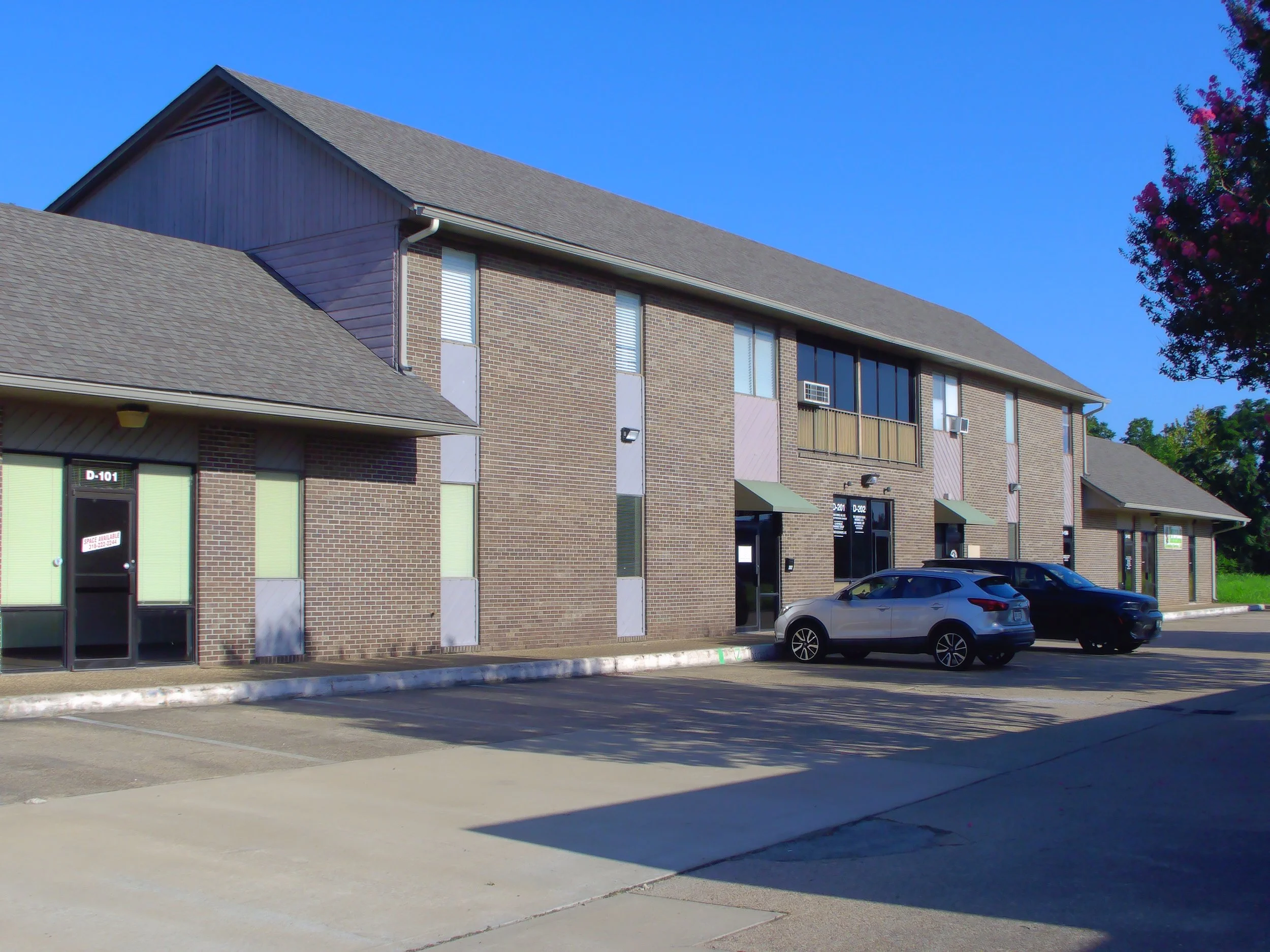 A two-story brick apartment complex with several windows, some with air conditioning units, and metal awnings above entrance doors. There is a parking lot in front with a silver car parked, and a tree with purple flowers on the right side. The sky is