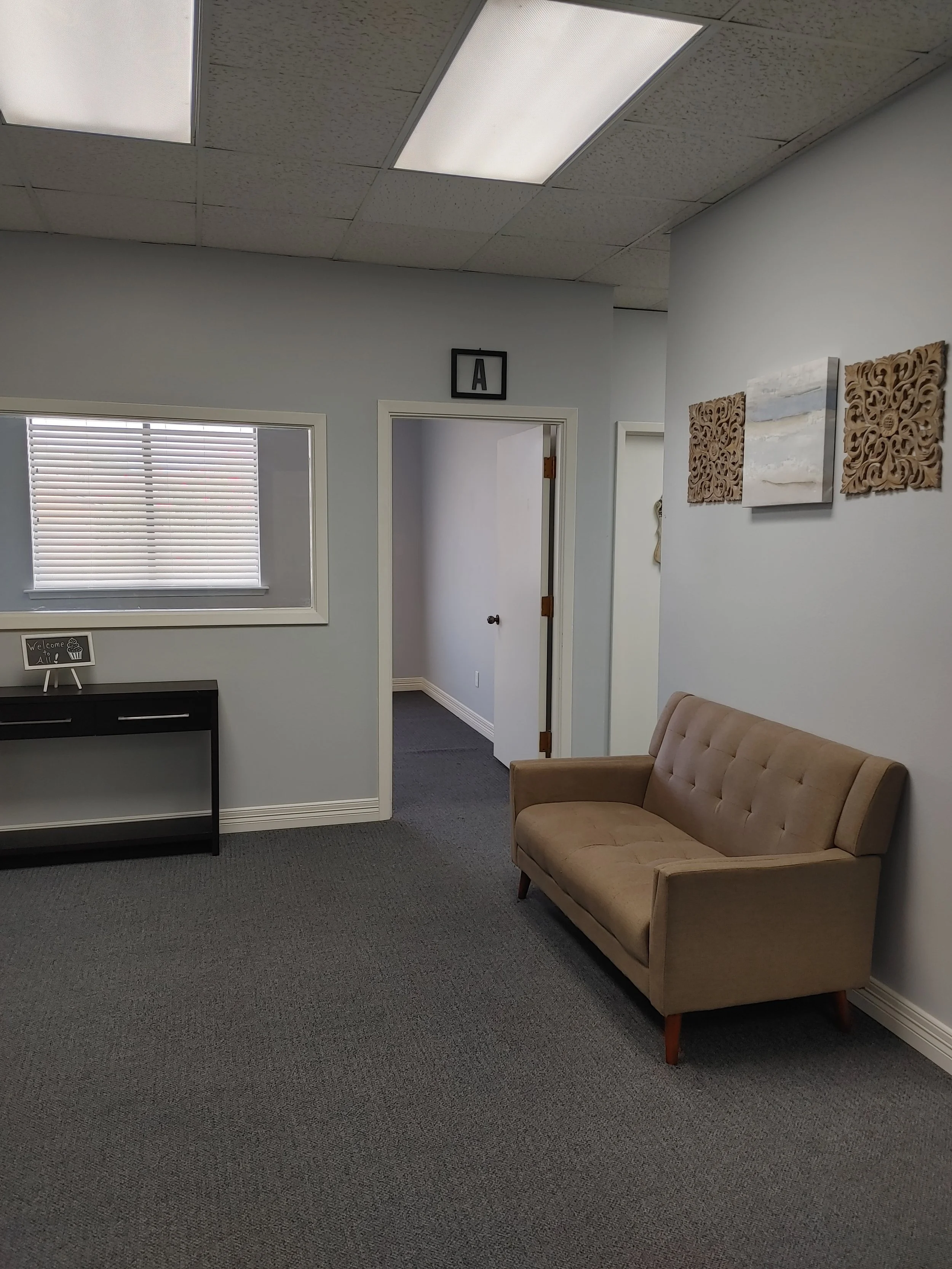 Office waiting area with a beige tufted sofa, a black desk with a small sign that says "Welcome All" and a window with blinds. The walls are painted light gray and decorated with abstract art. The ceiling has fluorescent lighting with textured tiles,