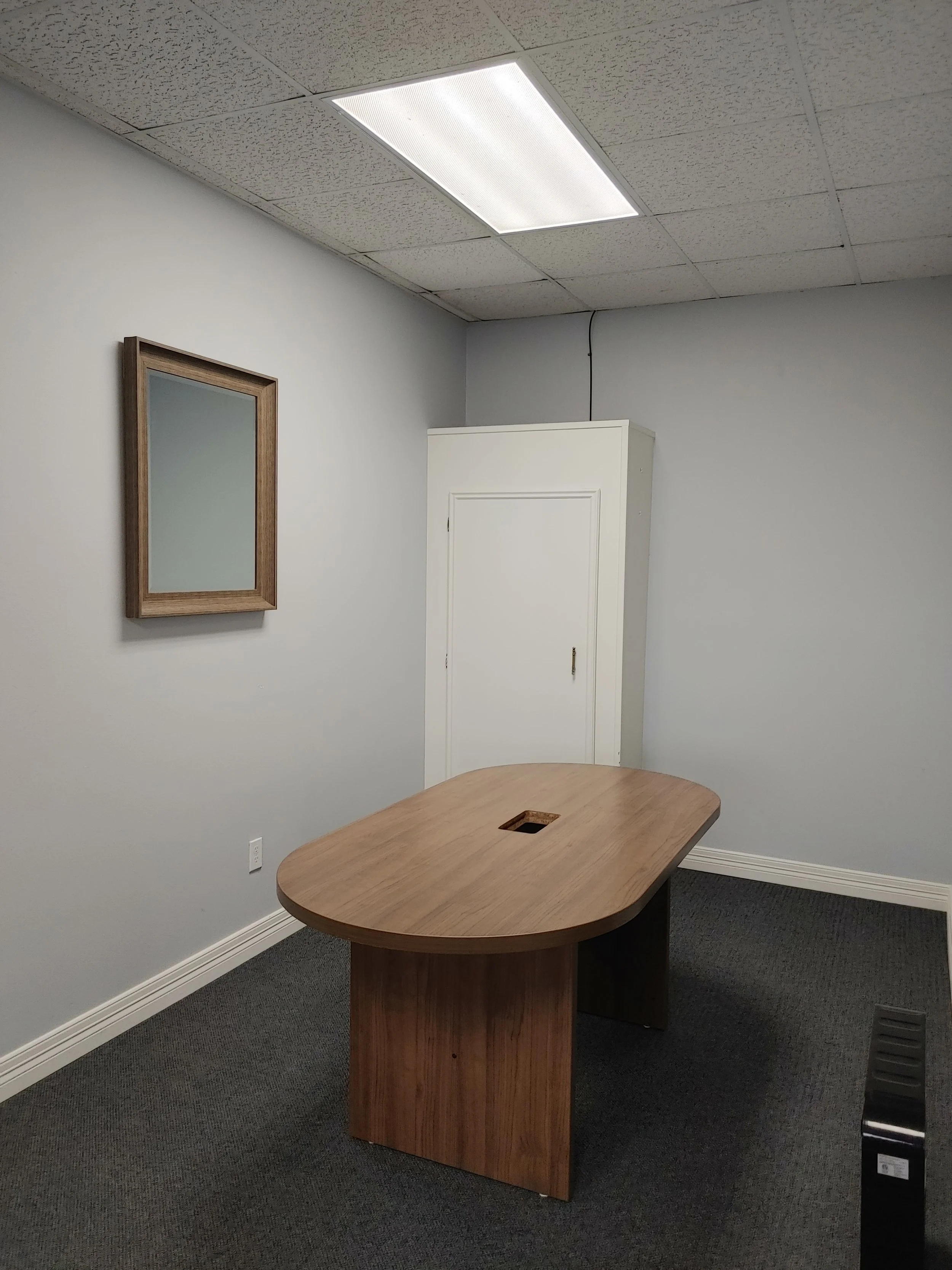 Office room with a wooden table, a mirror on the wall, white walls, a white door, ceiling tiles, and a fluorescent light fixture.