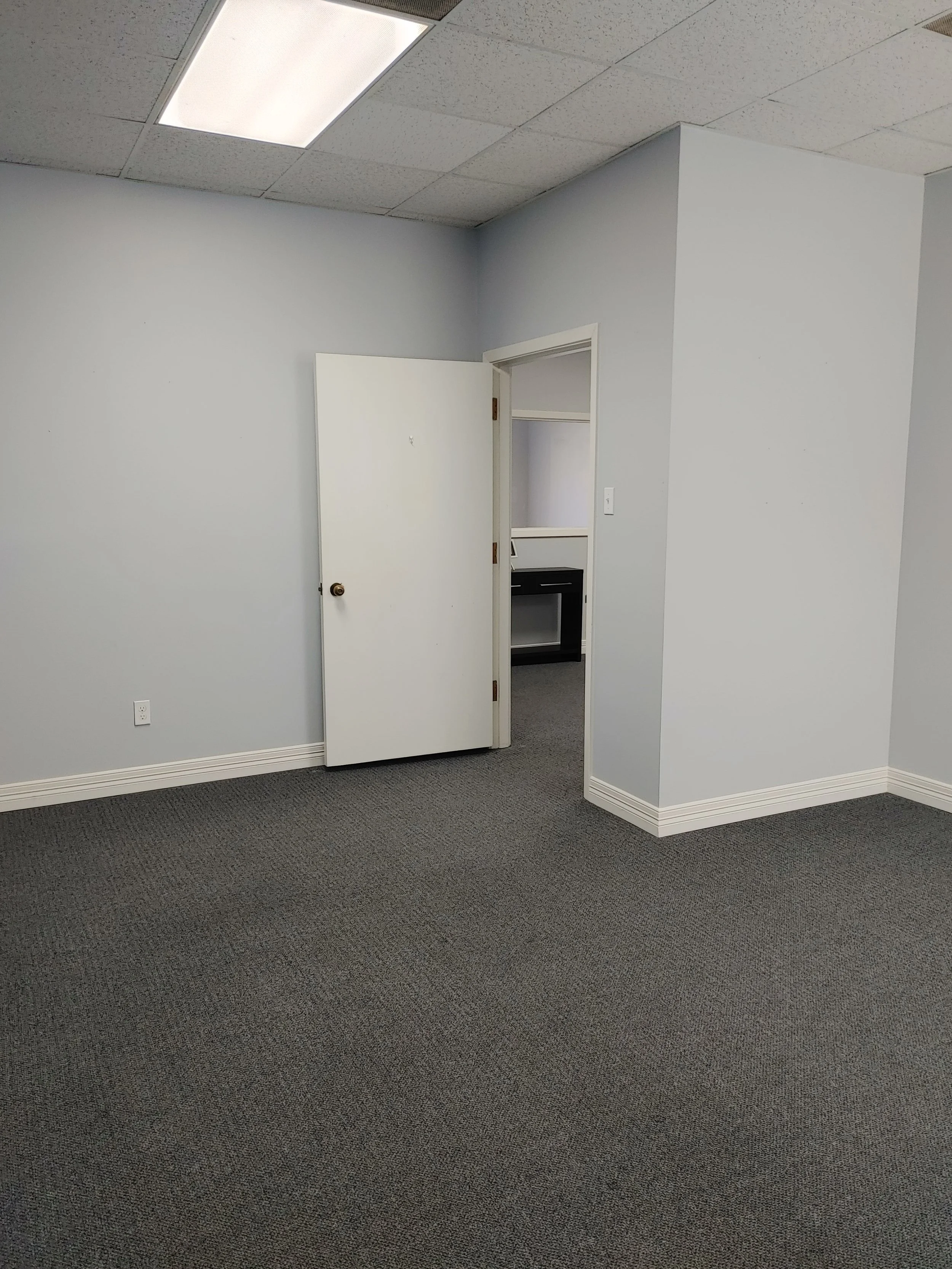 Empty office space with gray walls, gray carpet, a partially open door leading to a small kitchenette or utility area, and a ceiling with fluorescent lighting.