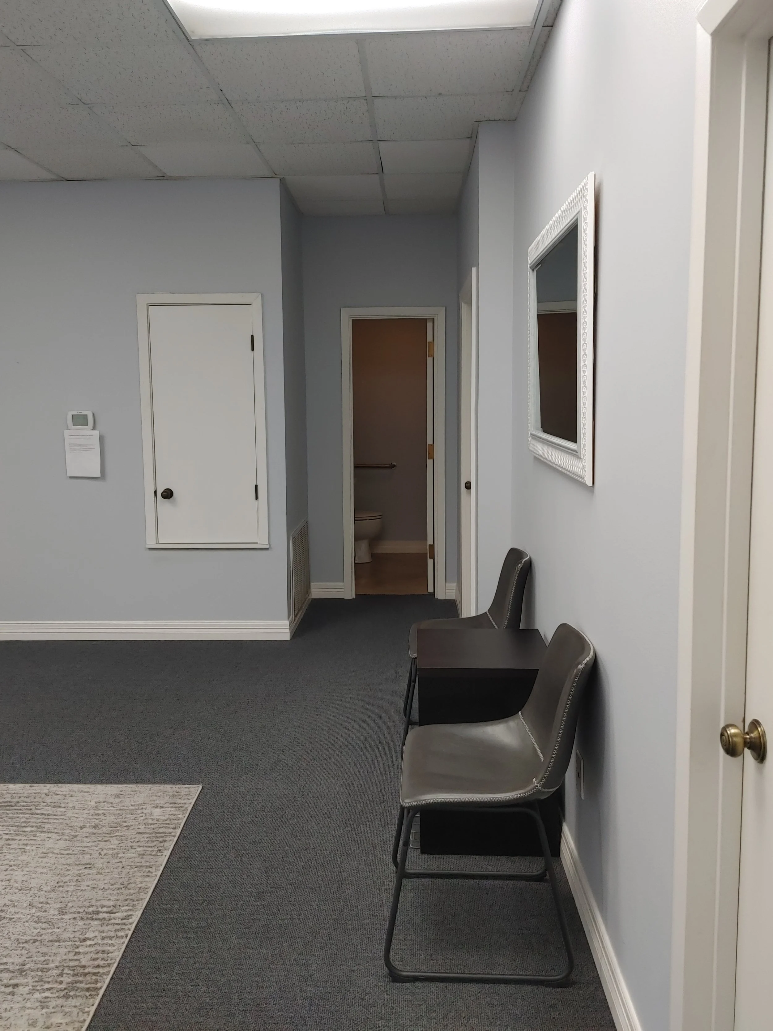 Interior of a waiting room with gray chairs, gray carpet, light gray walls, doors leading to restrooms, a mirror on the wall, and a small area rug on the floor.