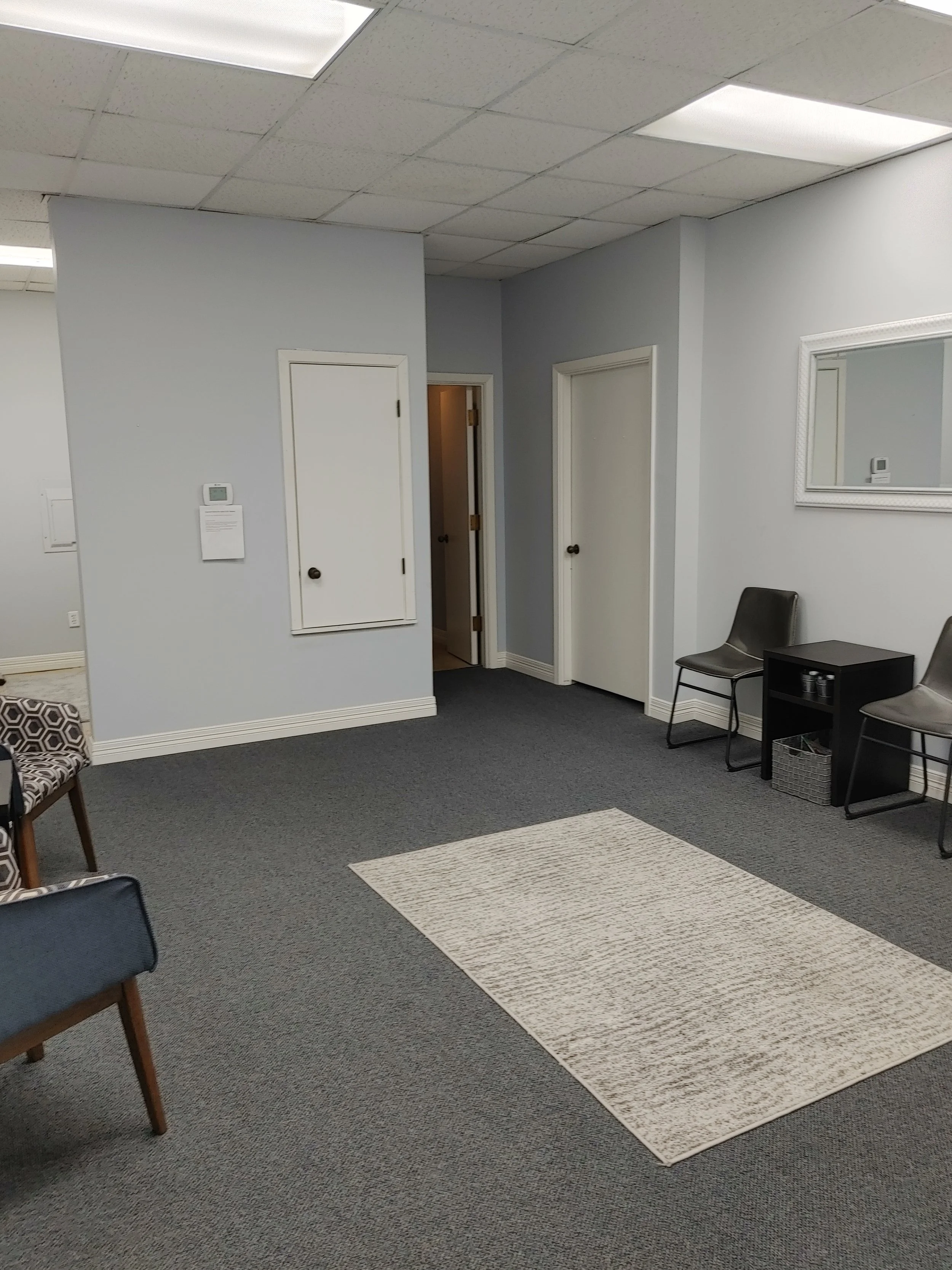 A waiting room with gray carpet, a beige rug, and chairs. The walls are gray with a mirror and a thermostat. There are three doors, one open, with fluorescent lights overhead.