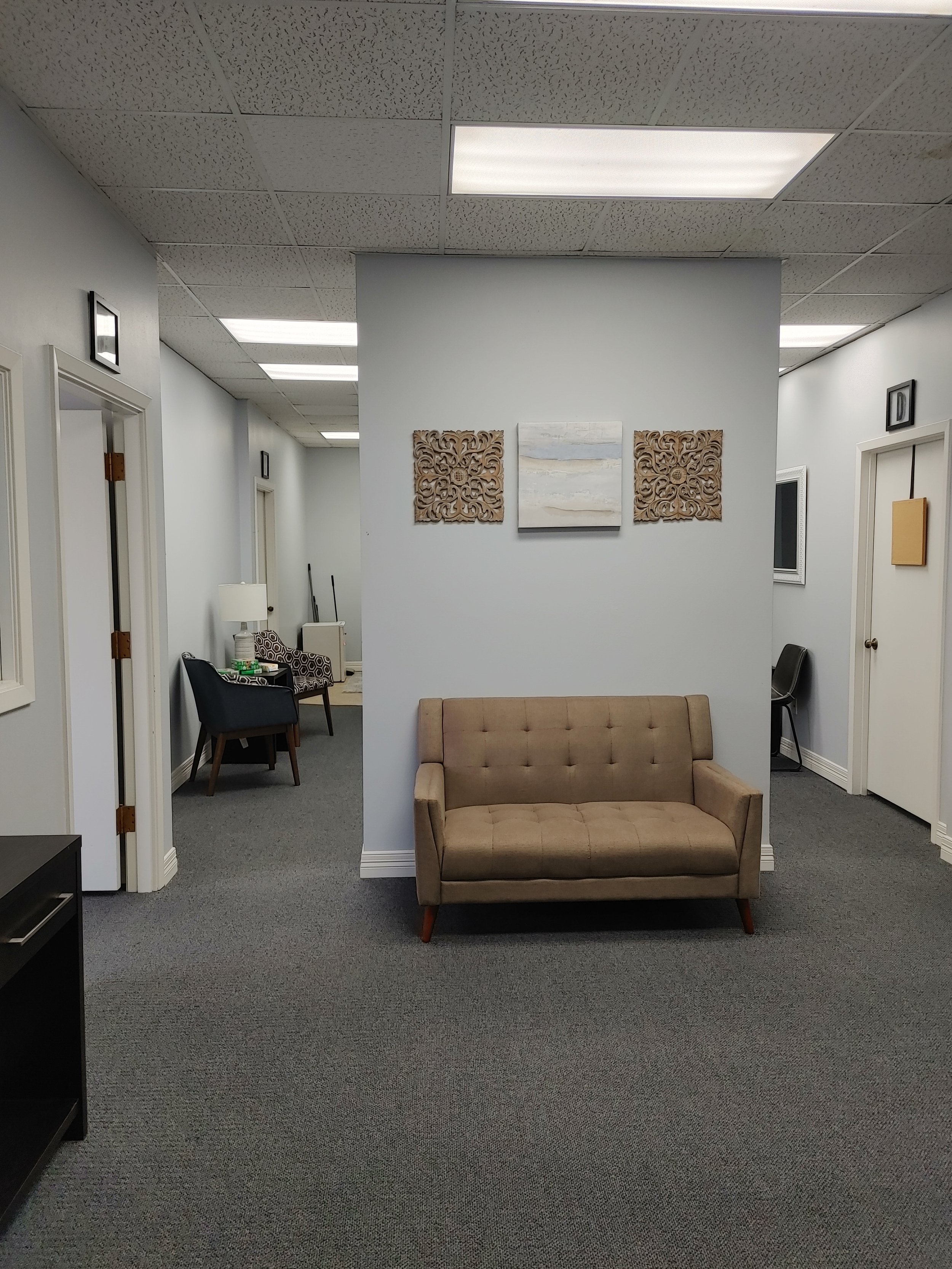 A waiting room with a beige sofa in the foreground, decorated with wall art and framed pictures, with chairs and small tables in the background, and a gray carpeted floor.