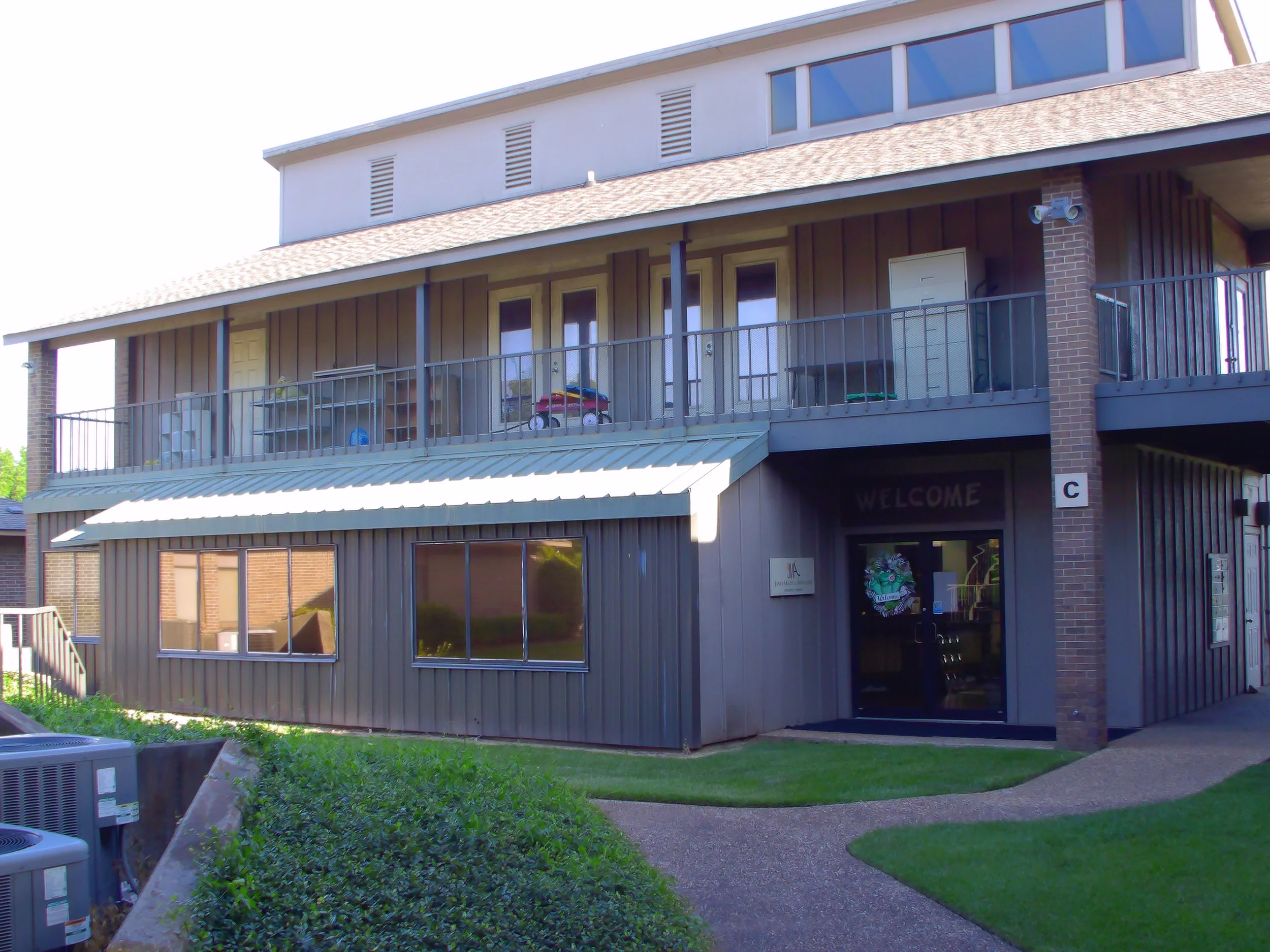 A multi-story building with a brick and beige panel facade, a balcony with a black metal railing, and large windows on the upper level. There is a welcome sign and some small items on the balcony.