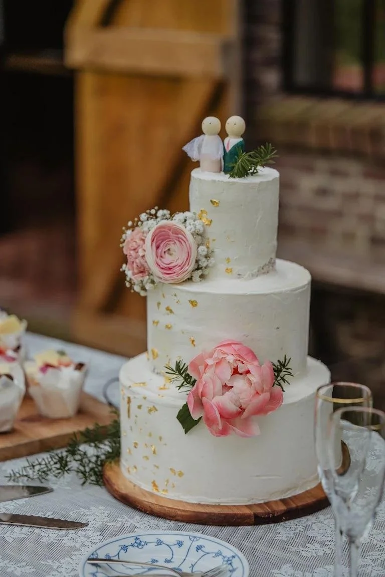 Drie-laags witte bruidstaart met bloemen en twee bruidspoppetjes als taarttopper, geserveerd op een houten plaat, met glazen en bestek op de tafel eromheen.