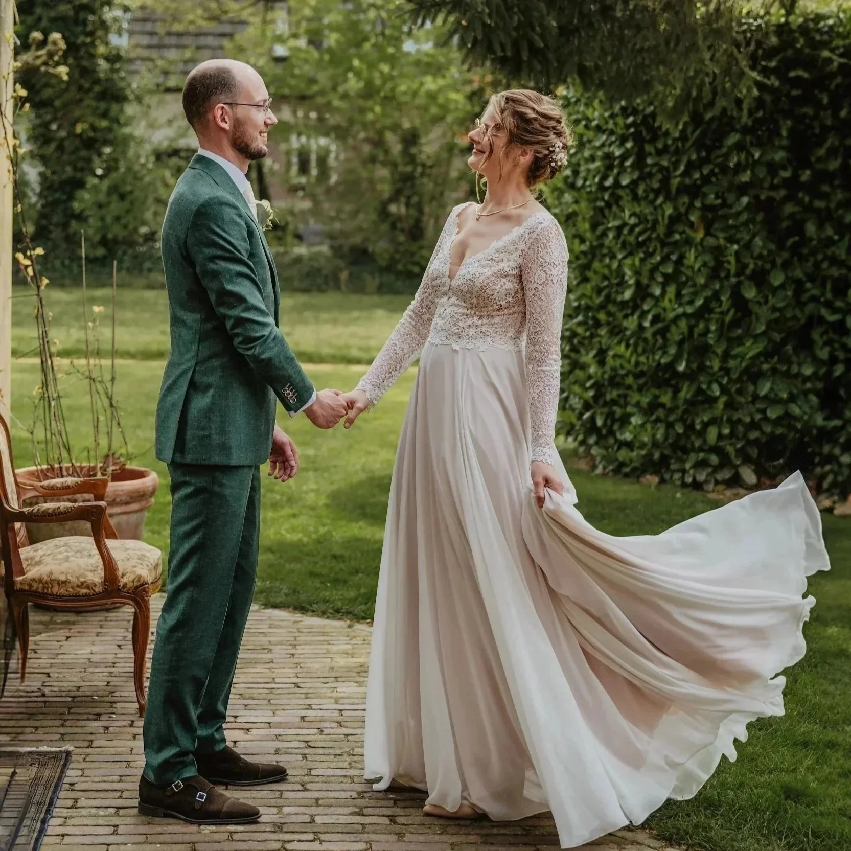 Een bruid en bruidegom die elkaar vasthouden en lachen tijdens een huwelijk in een tuin, met groen landschap en planten op de achtergrond.