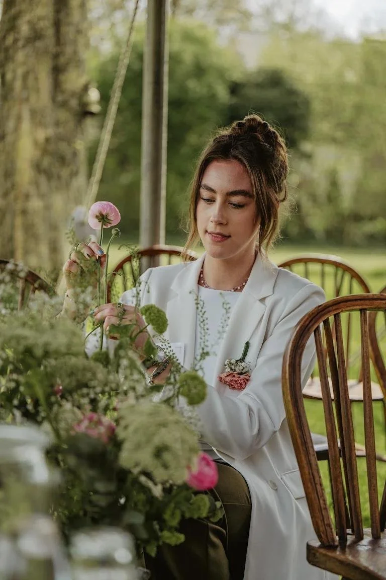 Vrouw die bloemen plukt in een tuin, omgeven door groene planten en ramen in een houten stoel.