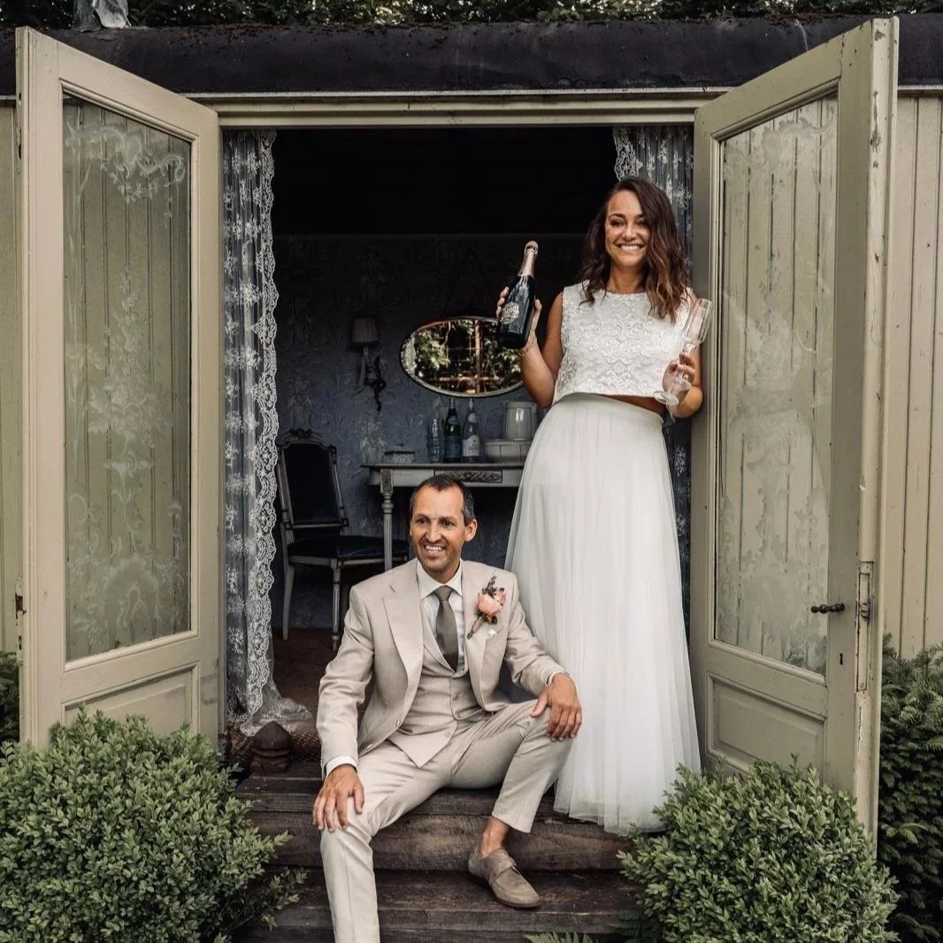 Een vrouw en een man, uitziend als bruid en bruidegom, poseren genietend op de drempel van een huis. De vrouw houdt een fles champagne en een glas, de man zit op de treden voor de deur.