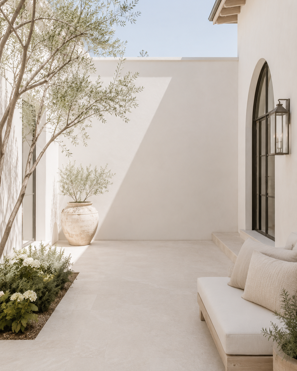 Bright Mediterranean-style courtyard with arched doors, olive trees, neutral stone, and outdoor seating.