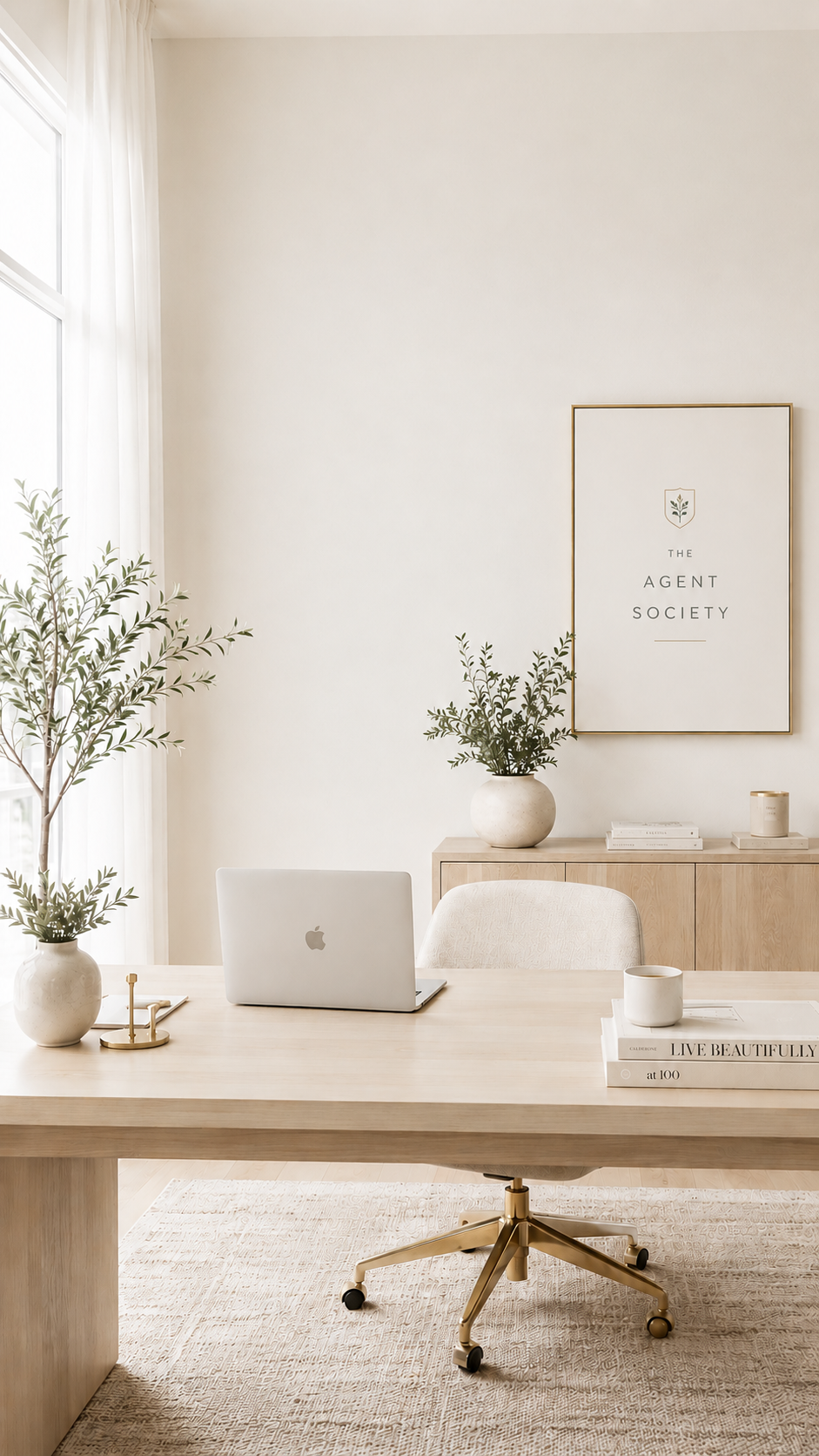 Bright, elegant real estate office with a light wood desk, laptop, greenery, coffee, and framed The Agent Society logo.