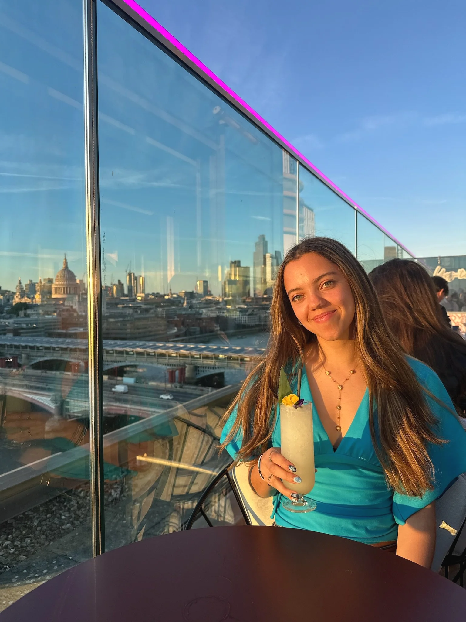 A young woman with long brown hair and green eyes holding a tropical drink in a tall glass with a flower garnish, standing next to a glass window with a city skyline in the background during sunset.
