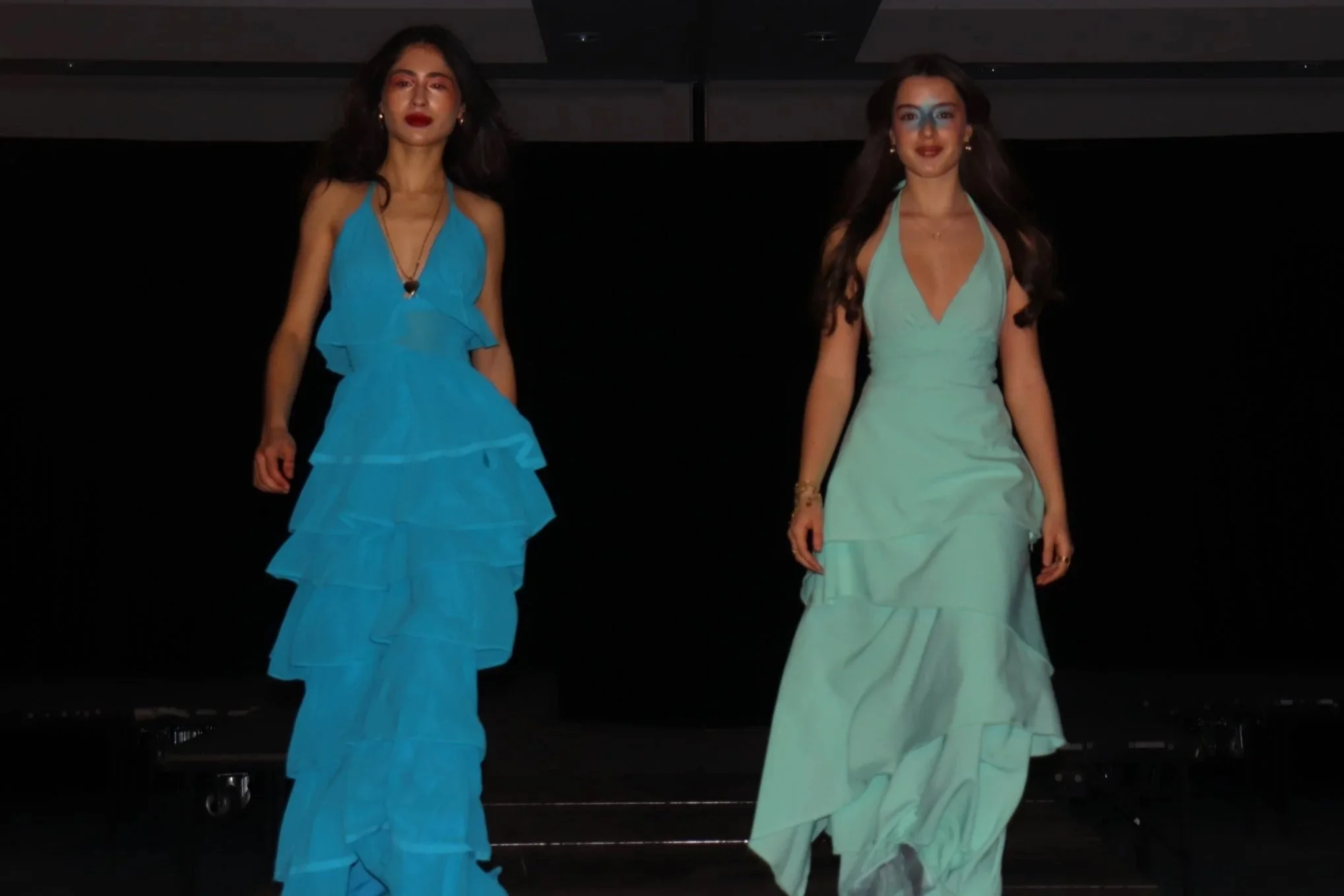 Two women walking on stage, each wearing a long, elegant dress; one in blue with ruffled layers and the other in light green with a halter neckline.