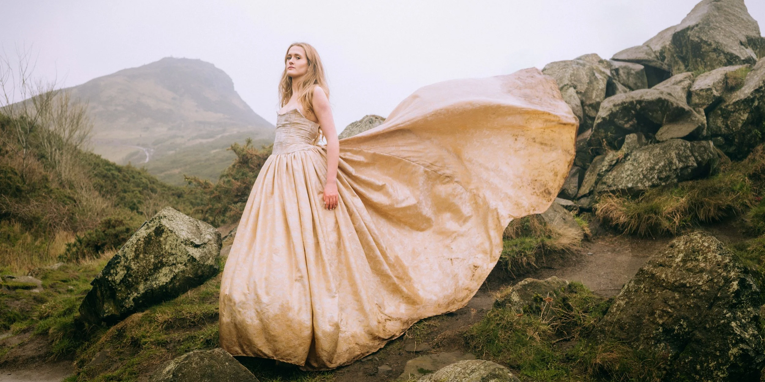 A woman in a flowing gold ball gown stands on a mountain among rocks and grass with mountains in the background.