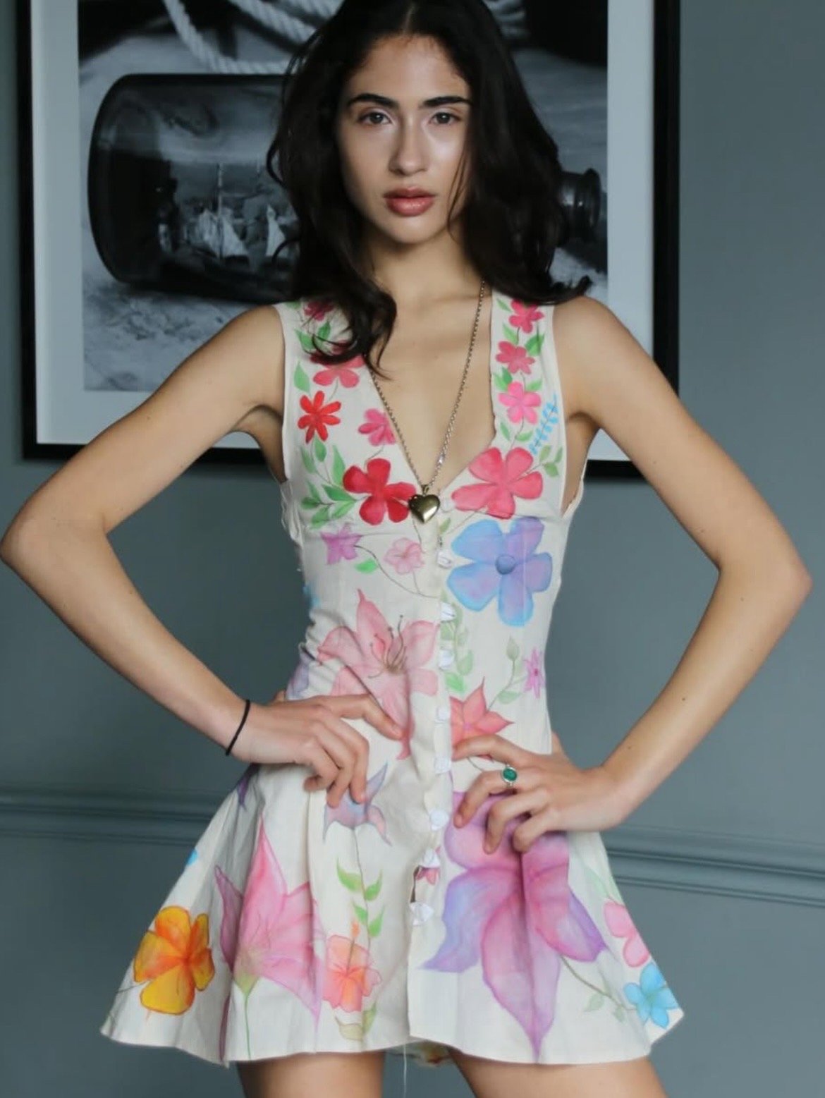 A young woman with dark wavy hair wearing a sleeveless floral dress with pink, purple, orange, and green flowers, standing with hands on hips in front of a gray wall with a framed black and white photograph of a fire extinguisher in the background.