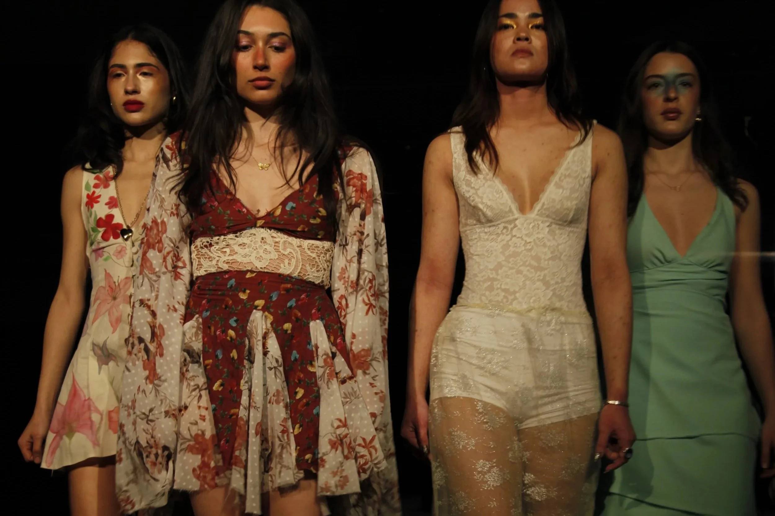 Four women standing close together in a dark setting, dressed in stylish outfits.