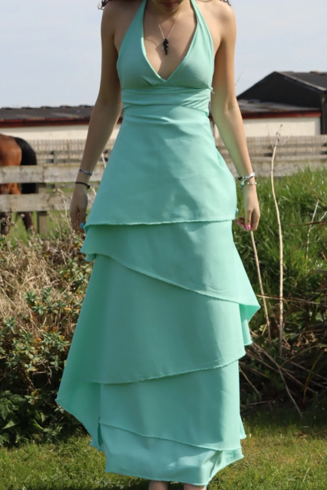 A woman wearing a mint green halter top and layered skirt standing outdoors in a grassy area, with a horse and wooden fence in the background.