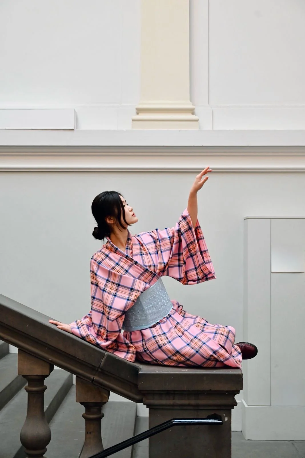 A woman dressed in a pink and blue traditional Japanese yukata is sitting on a staircase, pose gracefully with one hand raised and the other resting on the staircase, inside a building with white walls and decorative molding.