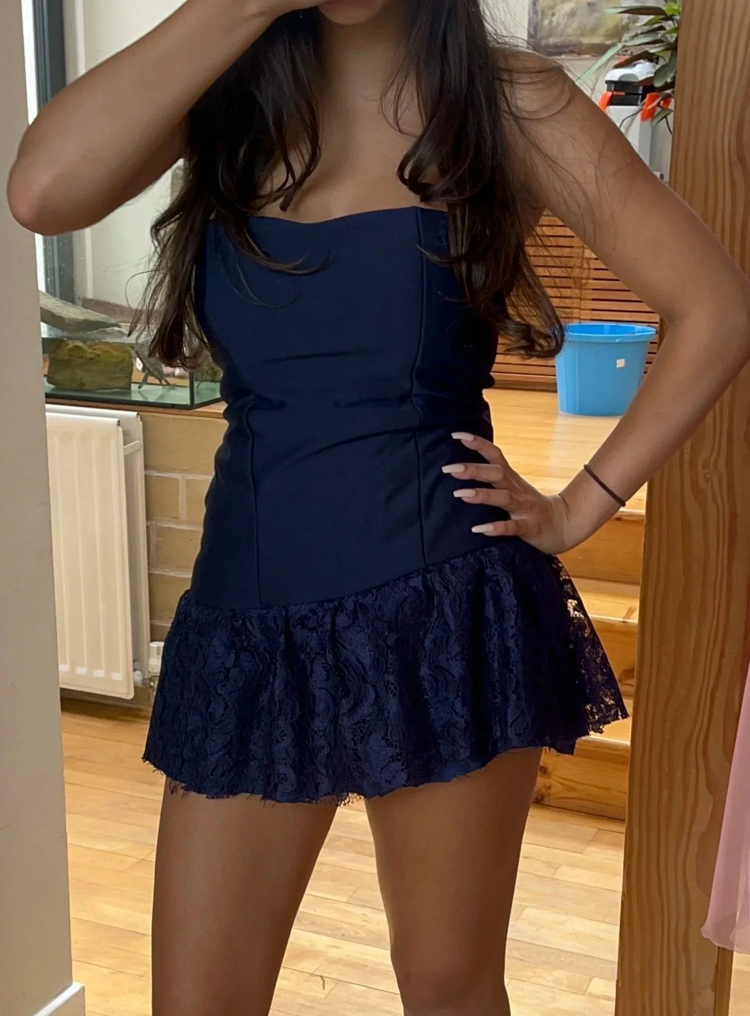 A woman taking a selfie in front of a mirror, wearing a dark blue strapless top and a lace skirt with her hand on her hip, in a room with wooden flooring and furniture.