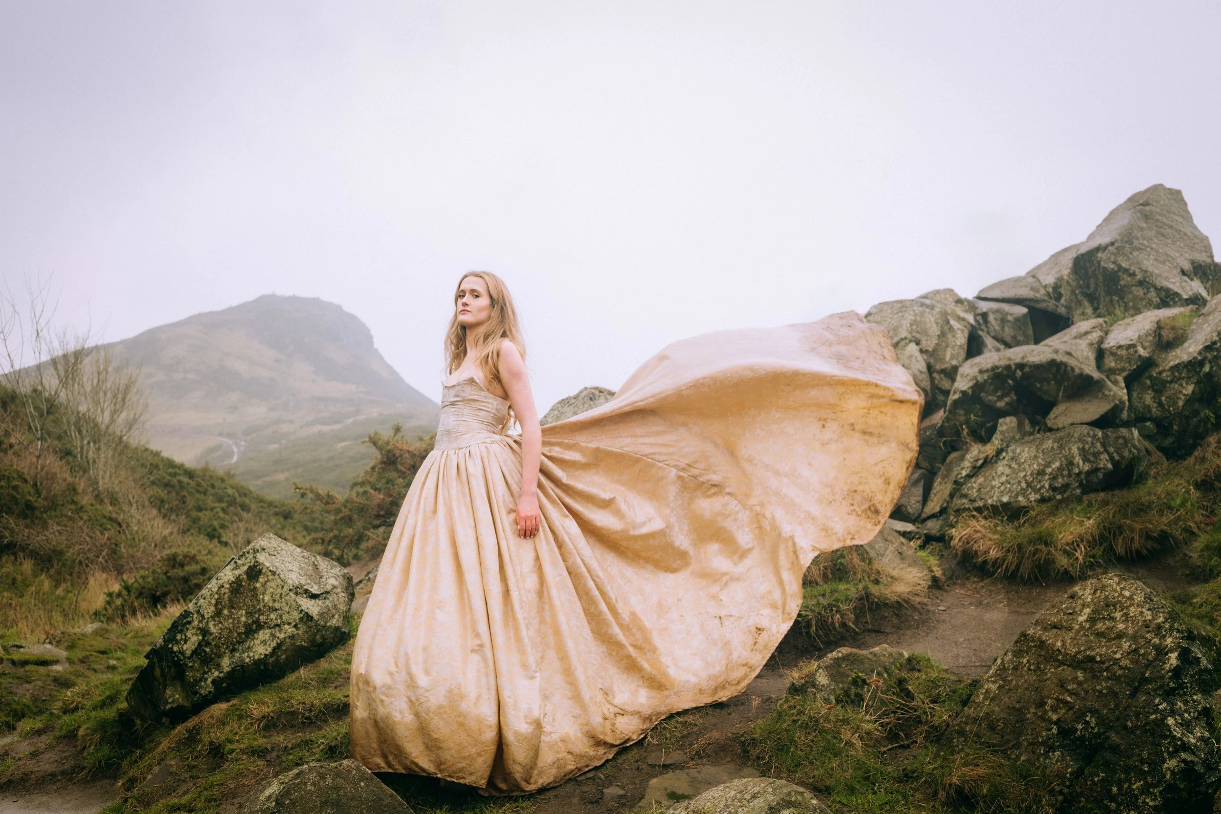 A woman in an elegant gold ball gown standing outdoors on rocky terrain with mountains in the background.
