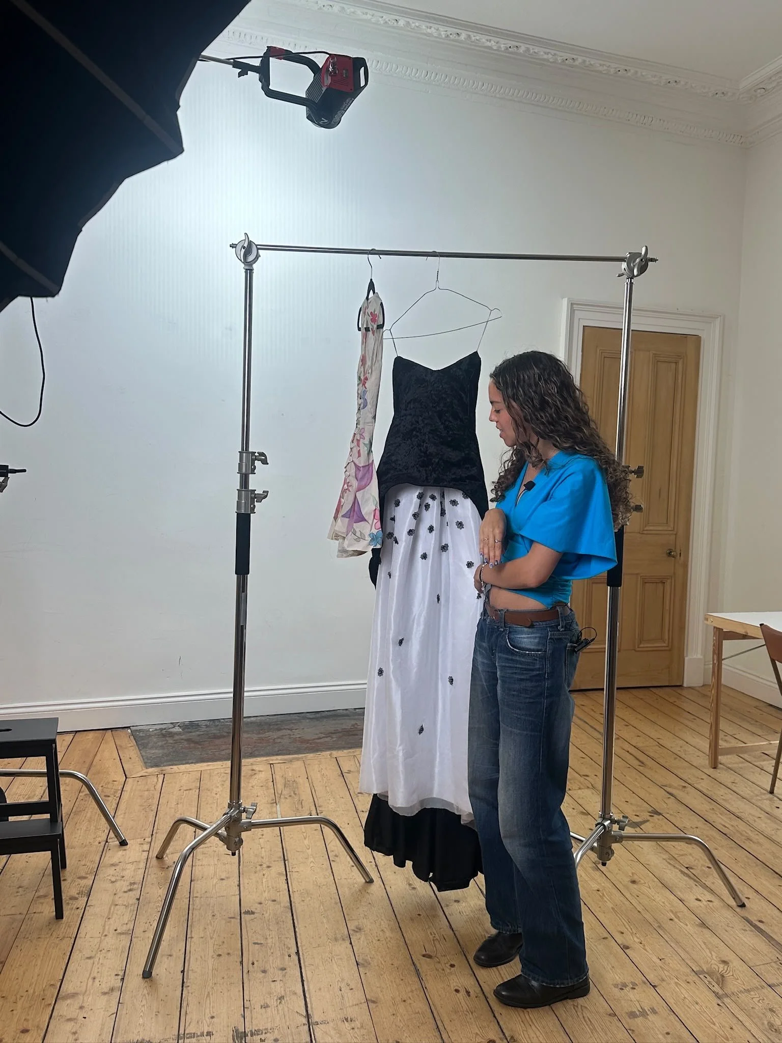 A woman in casual clothing stands with her eyes closed and hands clasped, in front of a dress form with a black top and a long white skirt with black patterns. The dress form has clothes hanging on it and is set up on a metal stand in a room with wooden floors and white walls, with photography lighting equipment surrounding her.