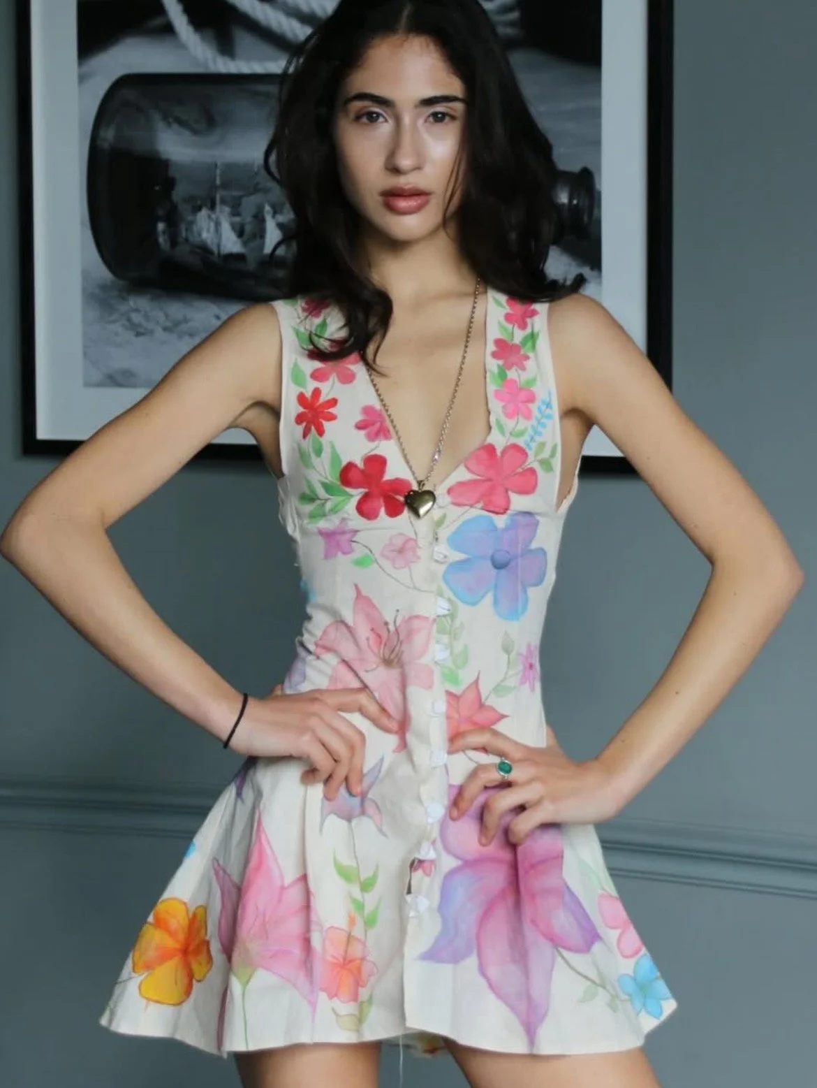 A young woman with dark hair, wearing a sleeveless floral dress with pink, blue, orange, and green flowers, standing with hands on hips in front of a gray wall and black-and-white framed photograph.