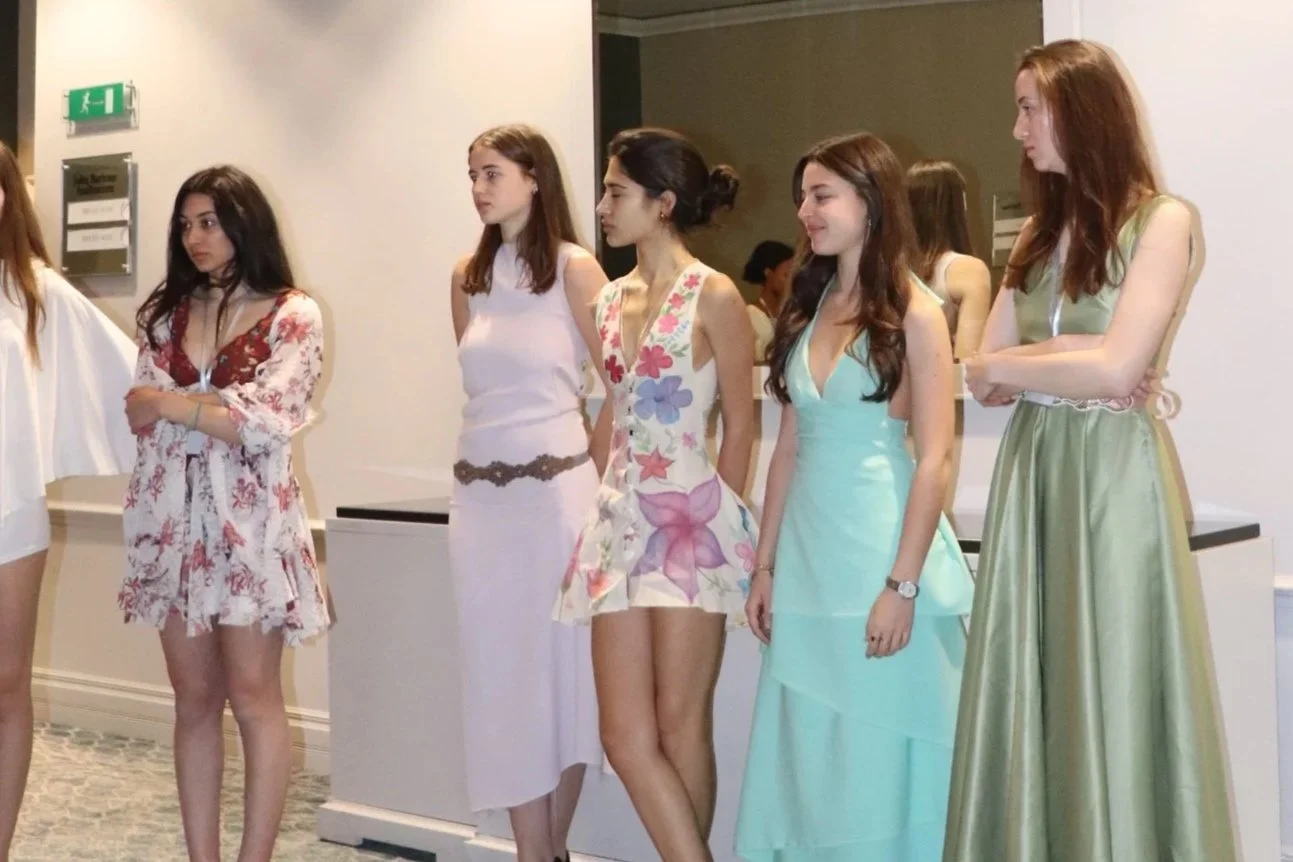 Six young women standing indoors, dressed in summer or formal dresses, with some crossing their arms and others standing with hands by their sides.