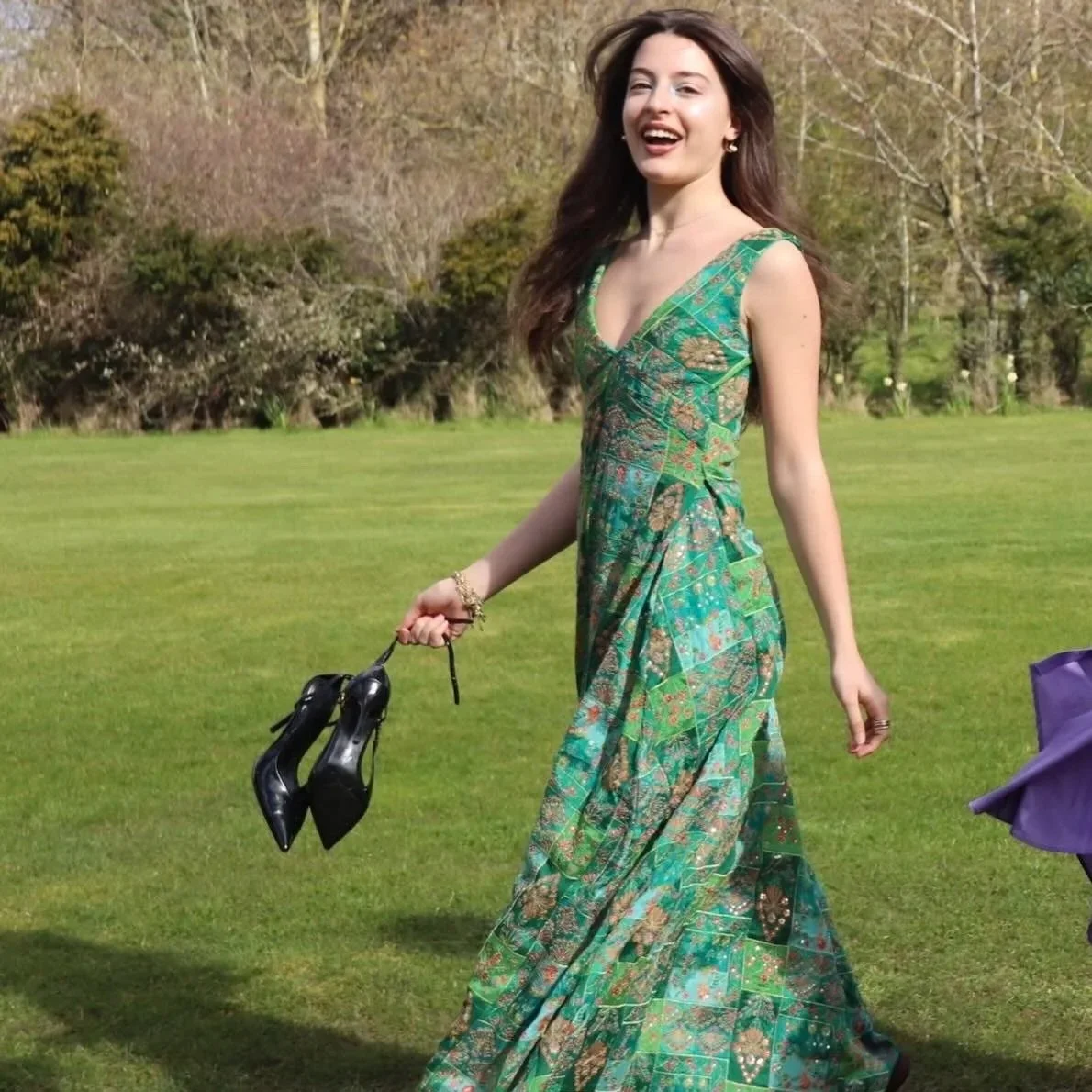 A woman with long brown hair wearing a green patterned dress is walking on a grassy field, holding a pair of black high heels in her right hand. She appears to be smiling or laughing.