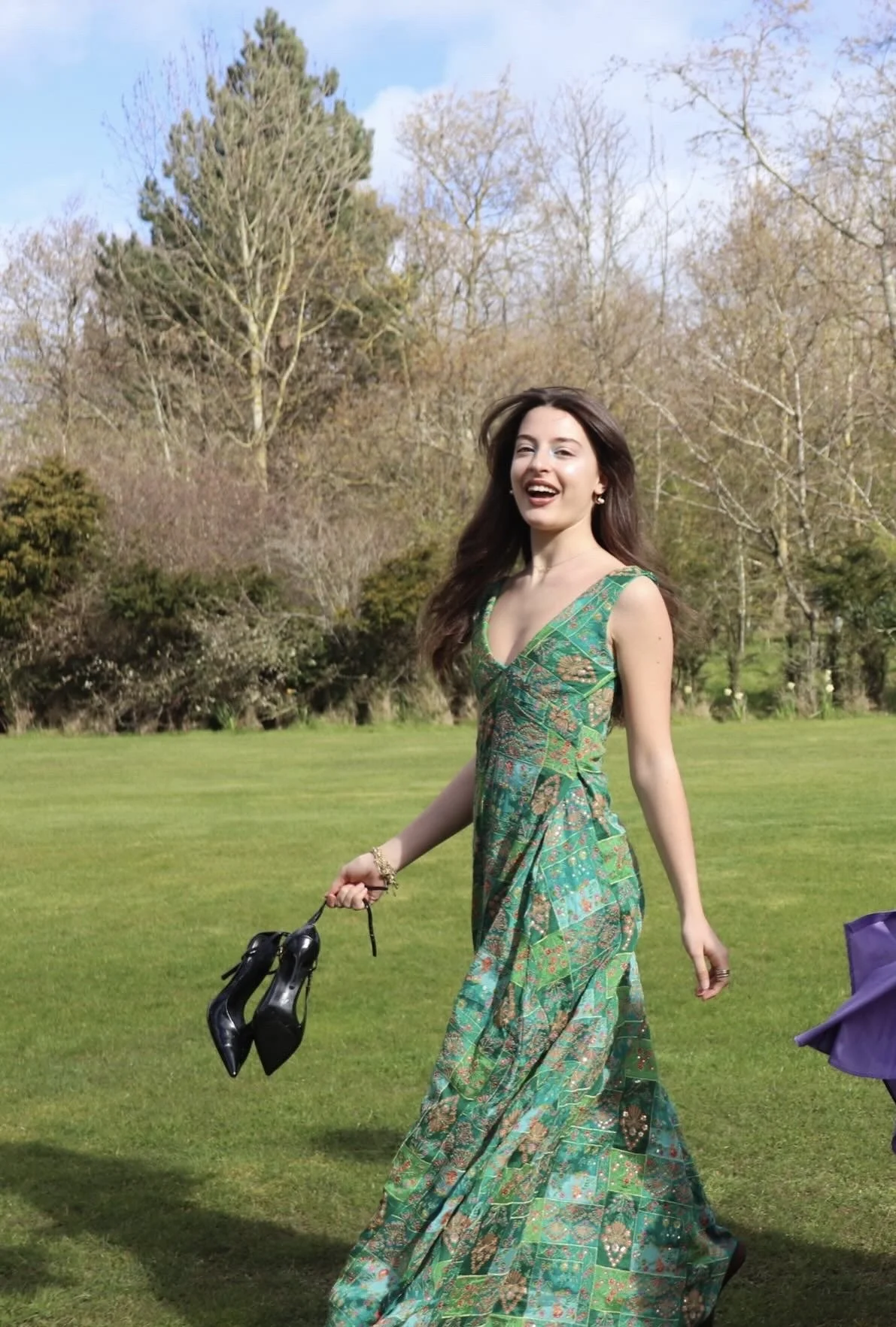 A woman in a long green patterned dress walking on a grassy field, holding a pair of black high heel shoes, with trees and a blue sky in the background.