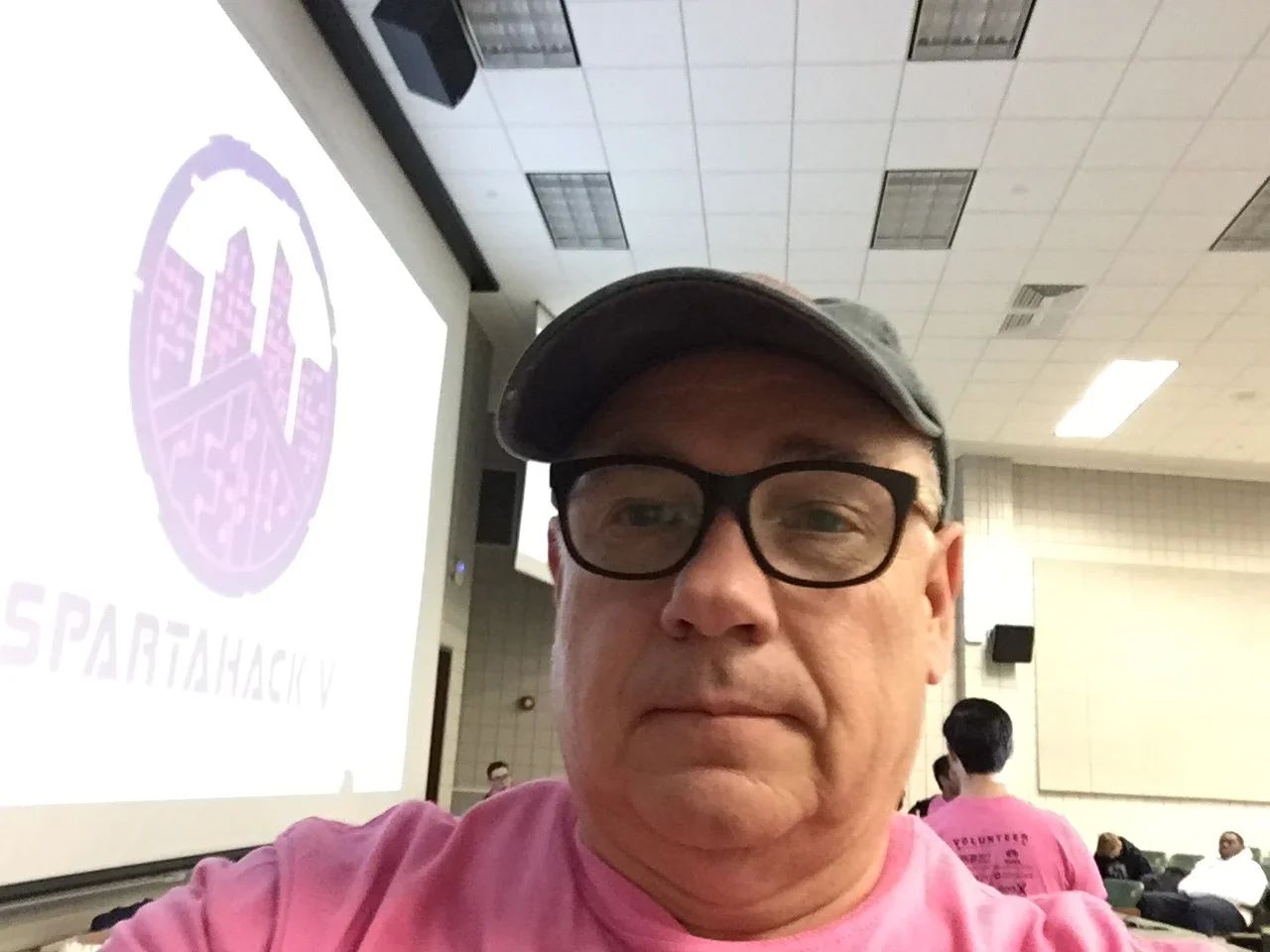 A man wearing glasses and a baseball cap taking a selfie in a large indoor room with a presentation screen displaying a purple Spartanhacky logo and text, and several people in pink shirts sitting and standing in the background.