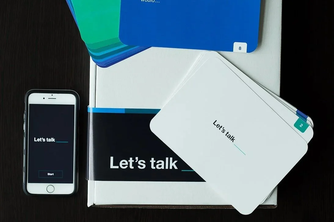 A smartphone displaying the words "Let's talk" with a button labeled "Start," placed next to a collection of cards and a book, all on a black surface.
