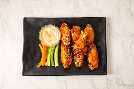 Plate with four chicken wings topped with sesame seeds, a small bowl of dipping sauce, and a few celery and carrot sticks on a black rectangular plate.