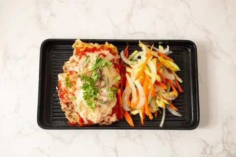 Baked chicken topped with mashed potatoes, cheese, and fresh herbs, served with mixed vegetable salad on a black tray.
