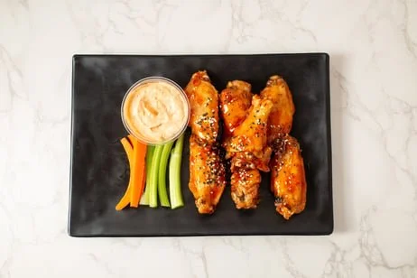 Four buffalo chicken wings with sesame seeds, a side of celery and carrot sticks, and a small dish of dipping sauce on a black rectangular plate.