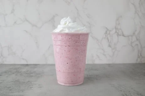A pink milkshake in a clear plastic cup topped with whipped cream on a gray surface with a marble background.