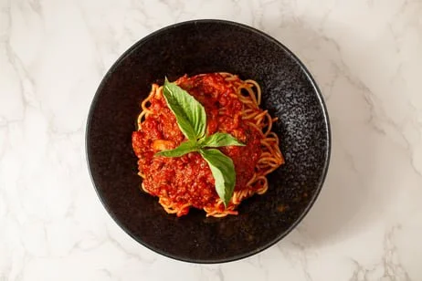 A black bowl of spaghetti with marinara sauce, garnished with fresh basil leaves, on a white marble surface.