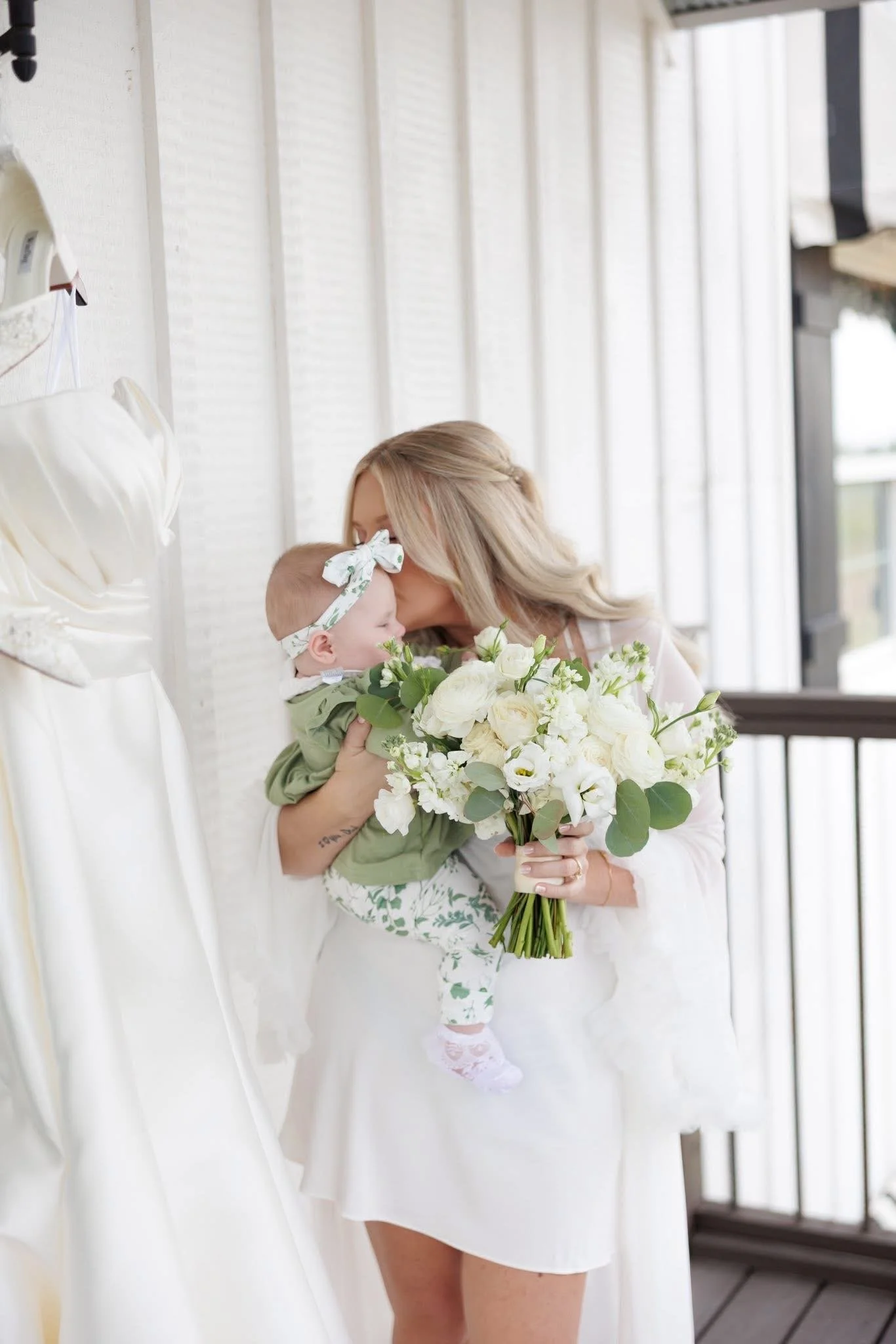 A woman holding a child with a bouquet of white and green flowers, standing in front of a white textured wall. The woman is kissing the child's forehead, and the child has a floral headband. The setting appears to be indoors near windows.