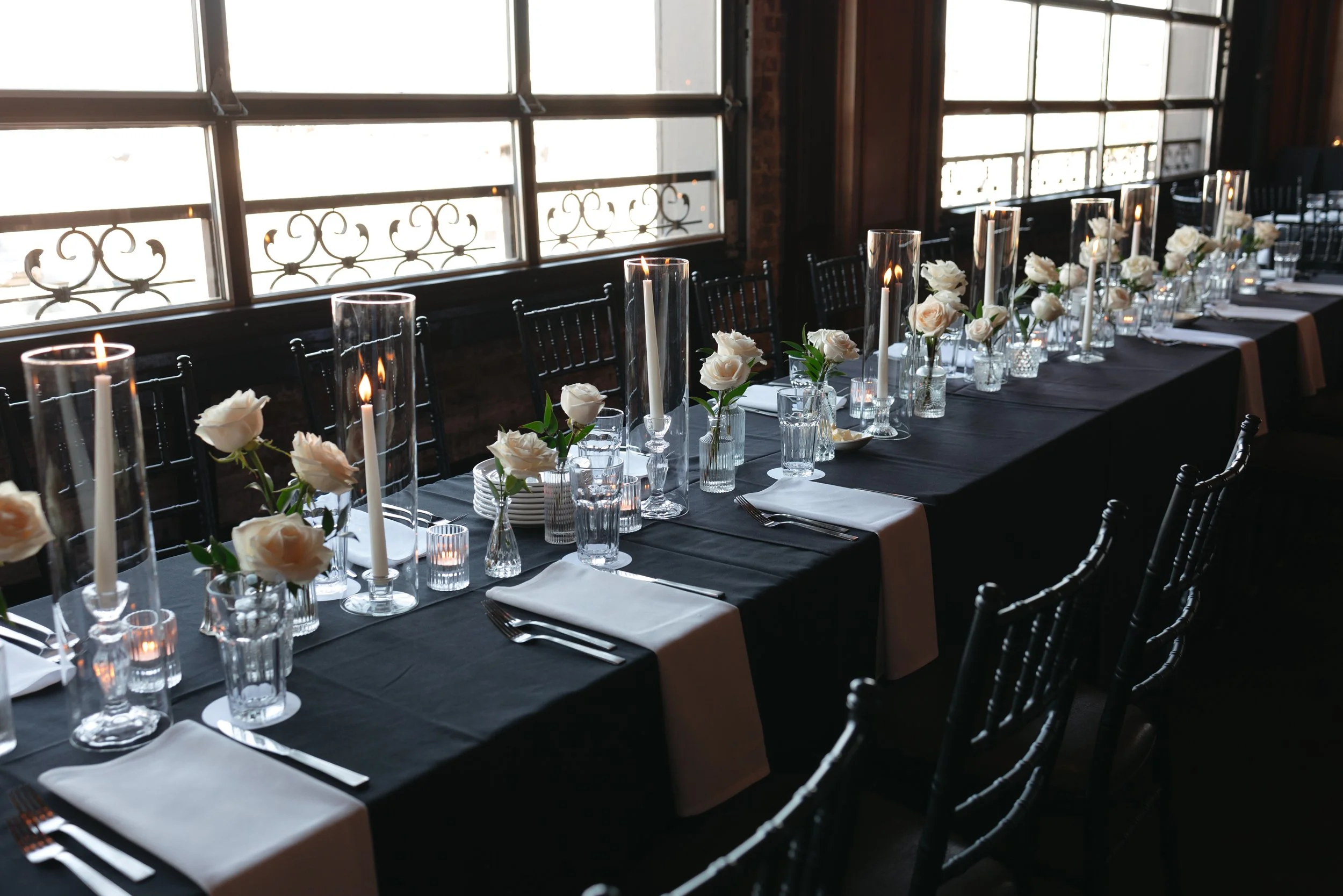 Long banquet table decorated with white roses in glass vases, tall white candles in glass holders, and table settings including white napkins and silverware, set near large windows with metal decorative railings.
