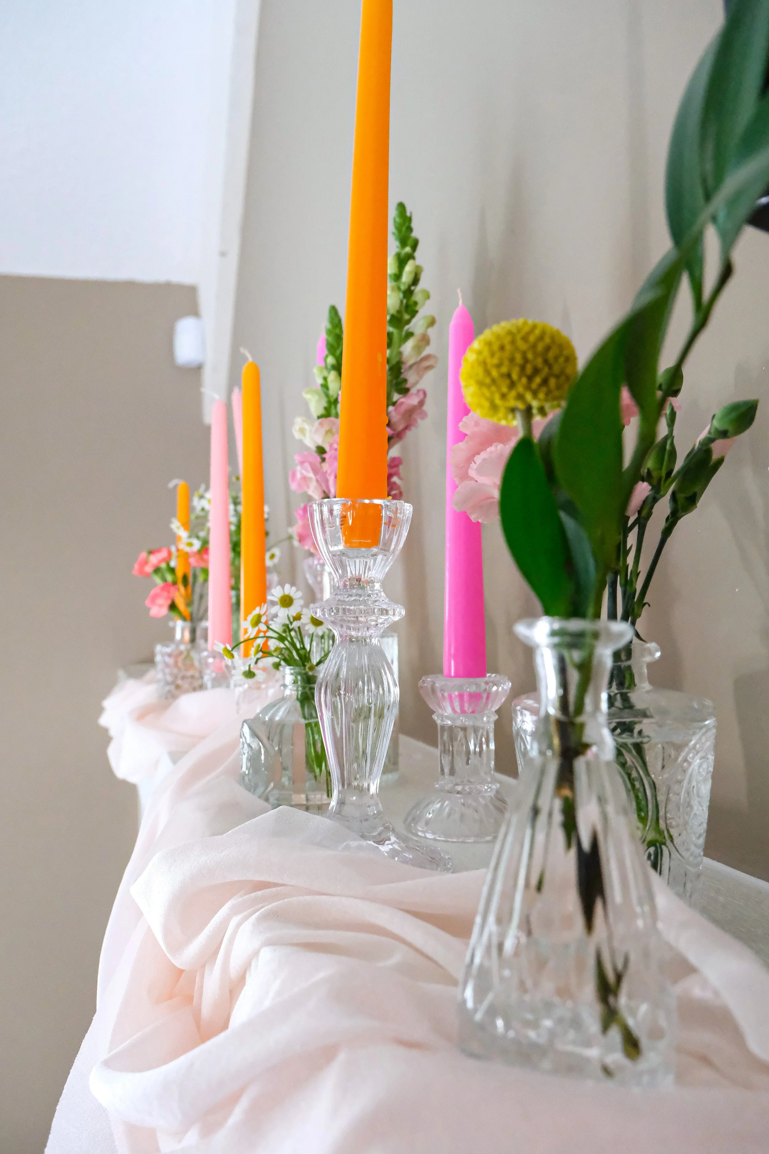 Close-up of a table with multiple glass candle holders, pink and orange candles, and assorted flowers, including yellow and pink blossoms, on a draped white cloth.