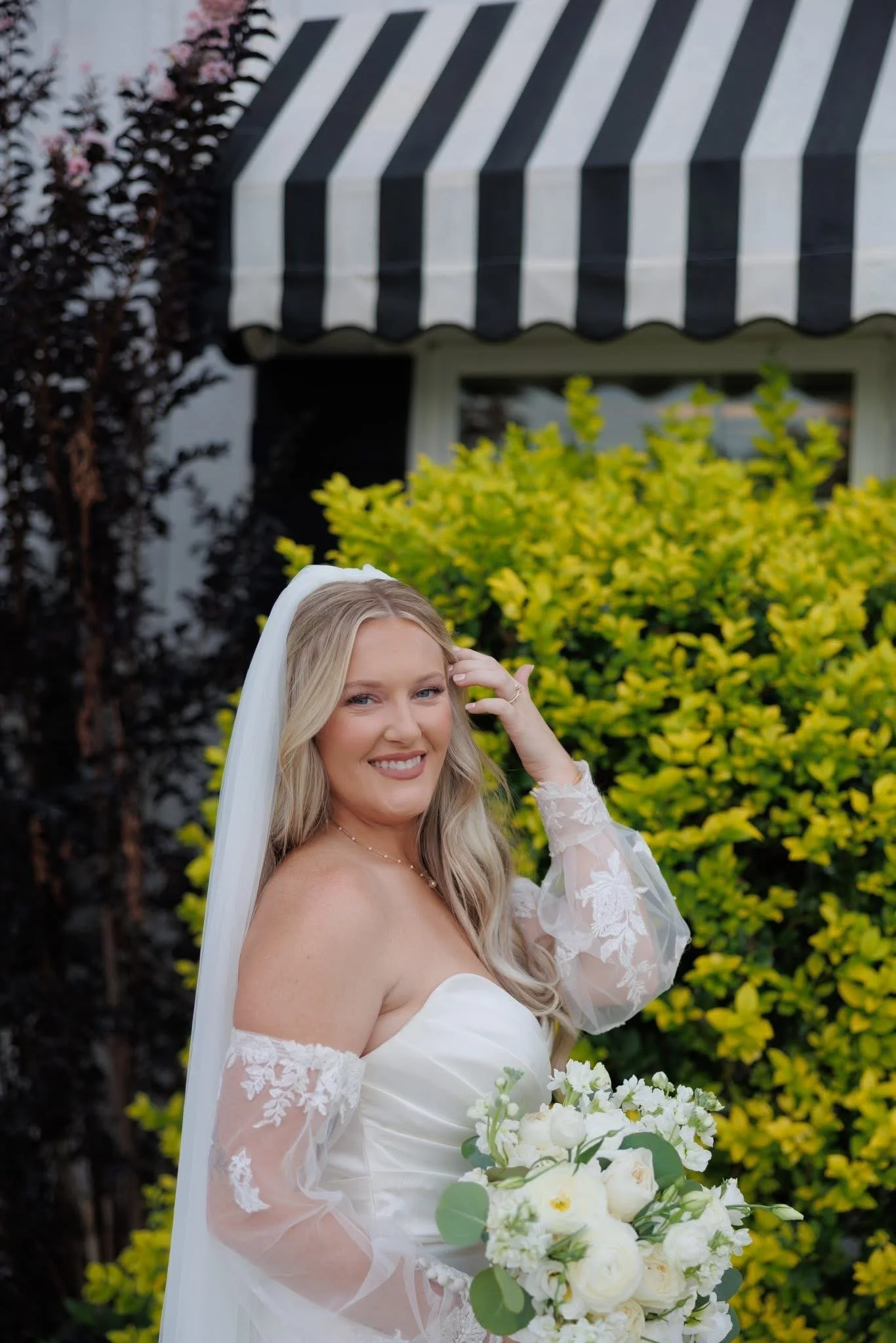 A woman in a wedding dress holding a bouquet of white roses and greenery, smiling outdoors with yellow and dark purple bushes and a black-and-white striped awning in the background.